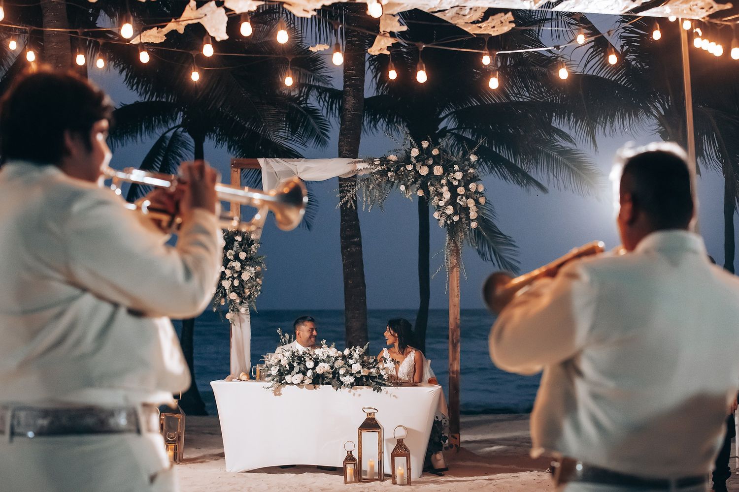 String lights illuminate an outdoor evening wedding reception with palm trees in the background.