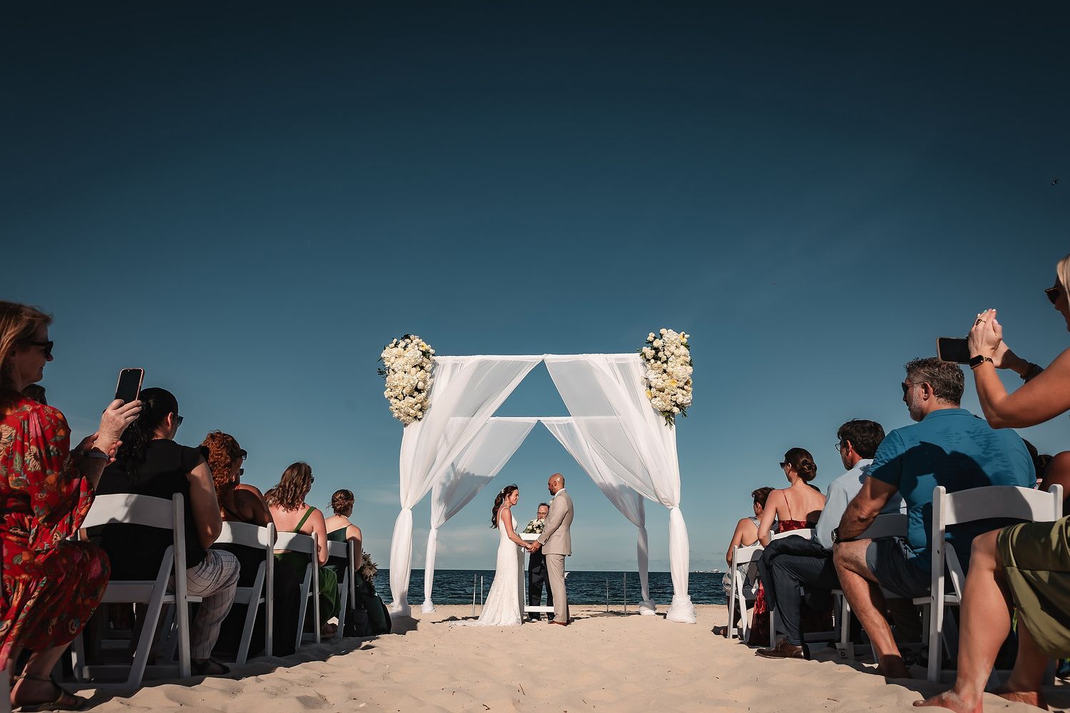 A beachfront wedding ceremony setup with white chairs and arch against blue ocean backdrop.