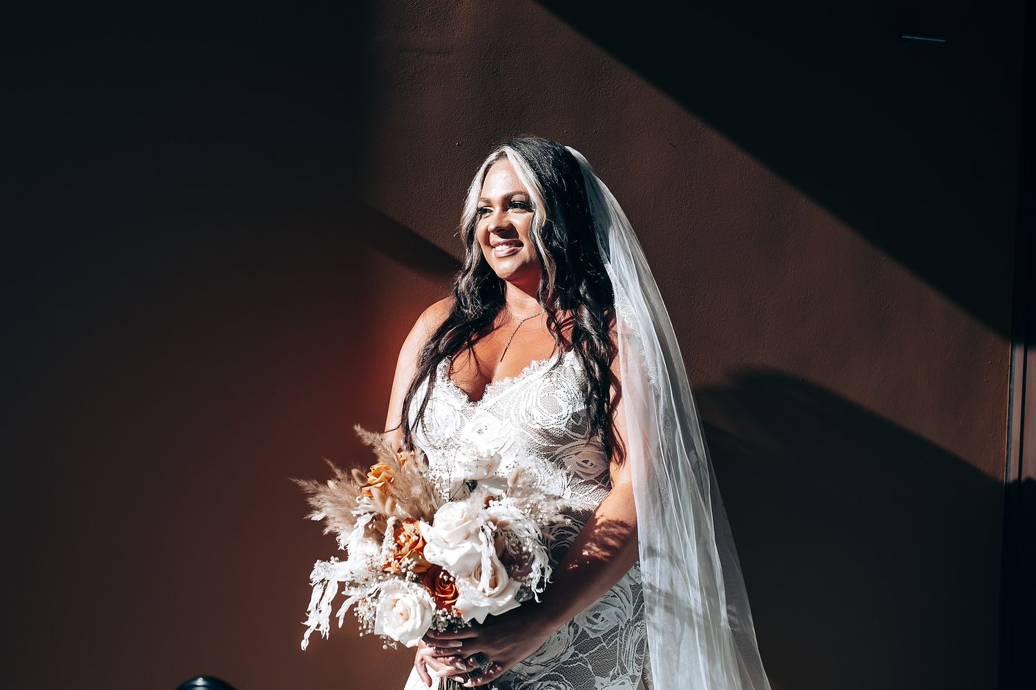 A bride in a white lace gown poses dramatically on a beach with palm trees and ocean views.