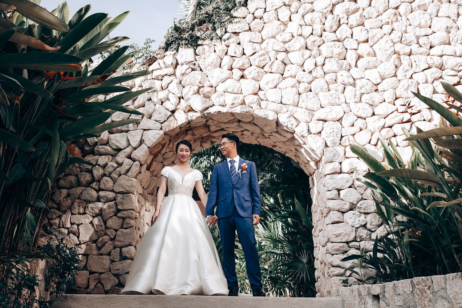 A couple poses on stone steps surrounded by lush tropical plants and a rustic stone wall backdrop.