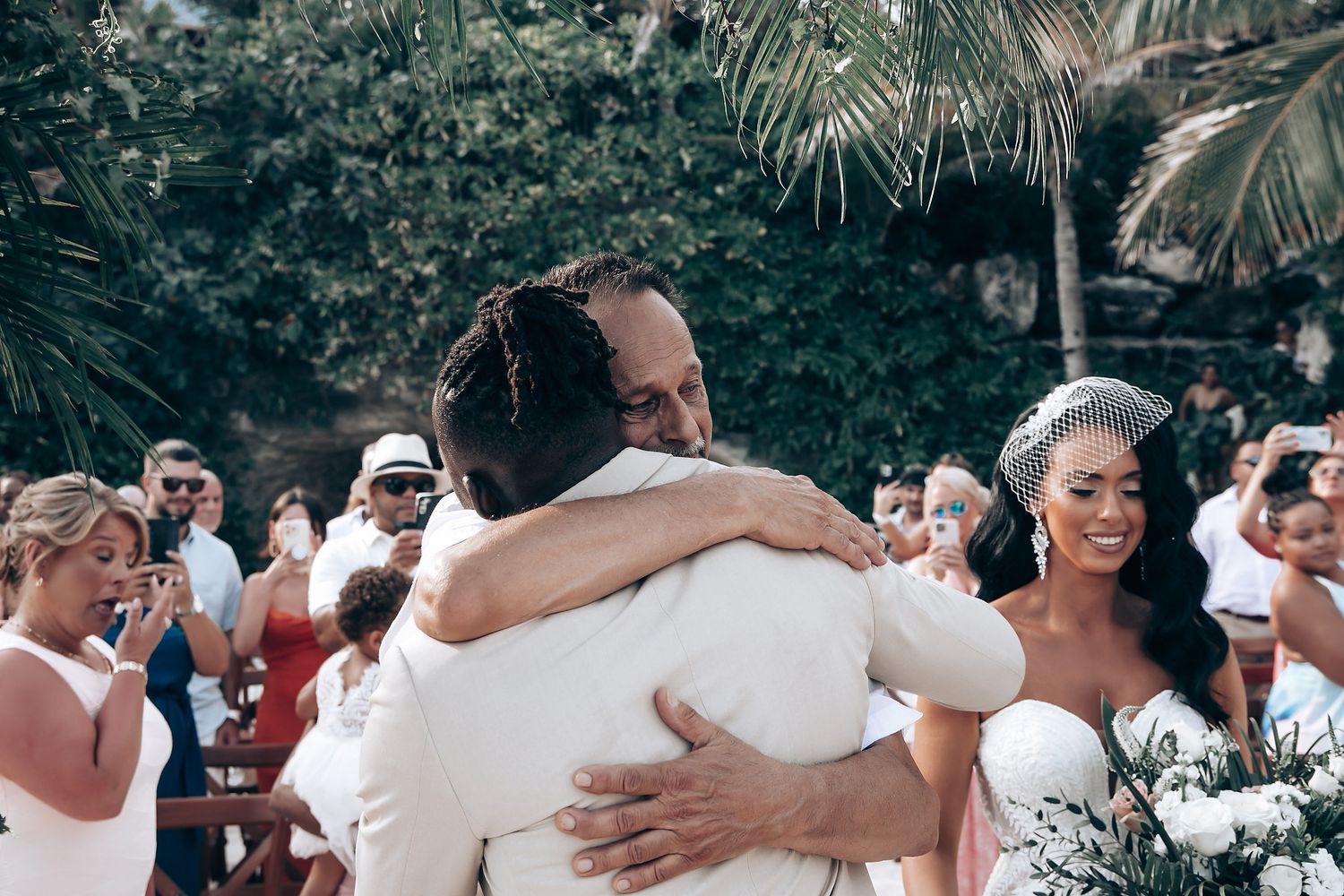 Wedding guests dressed in white embrace during an emotional group hug at an outdoor celebration.