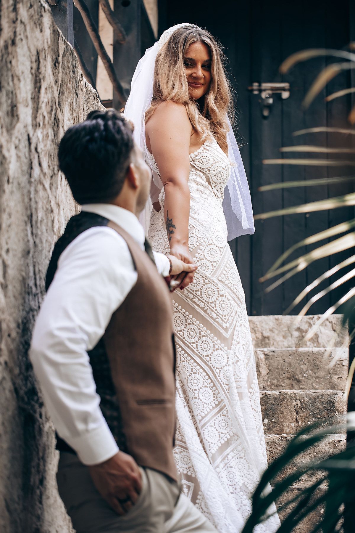 A couple in wedding attire share a romantic moment outdoors with palm trees and stone walls in the background.