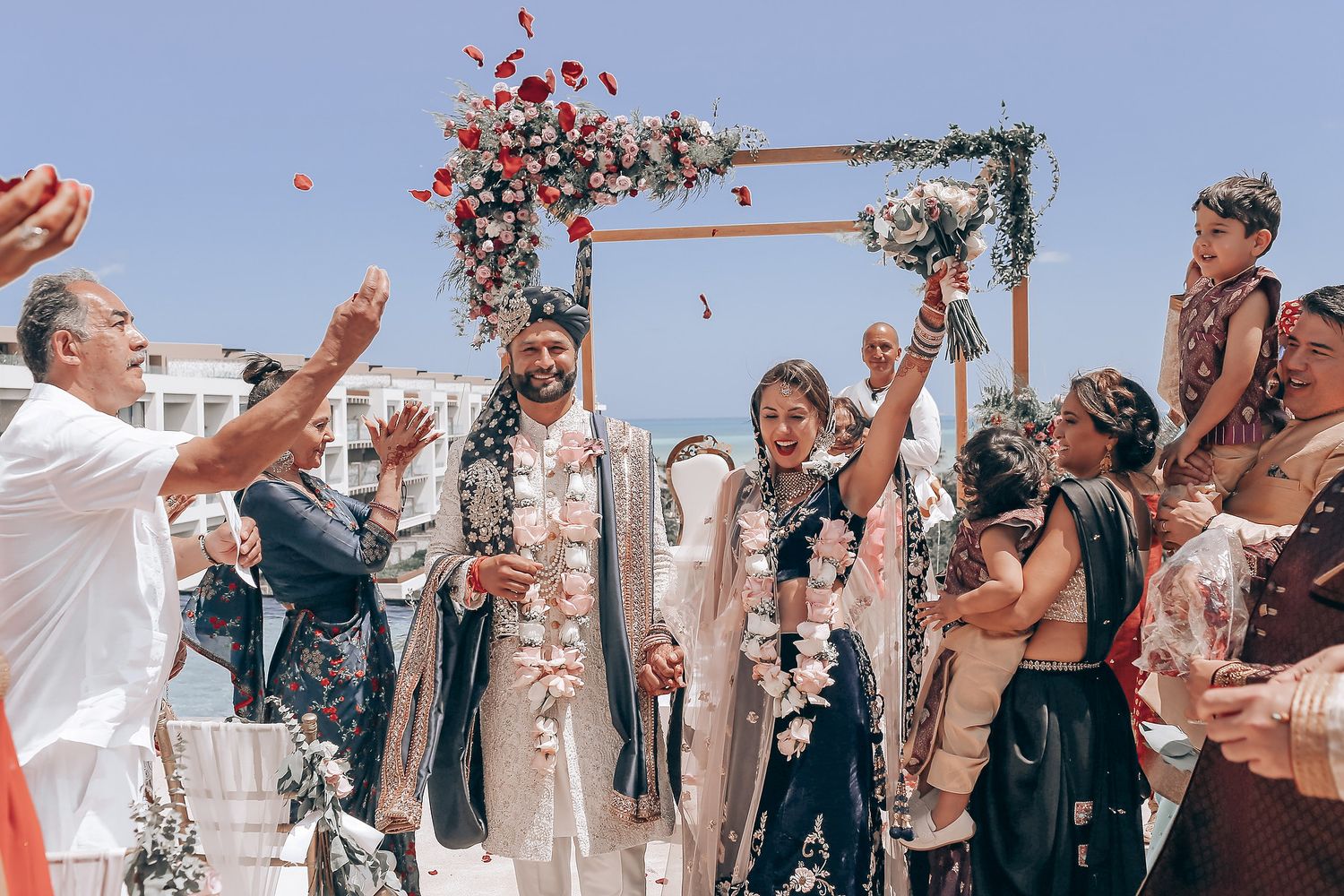 Wedding guests toss rose petals during an outdoor celebration on a sunny day.