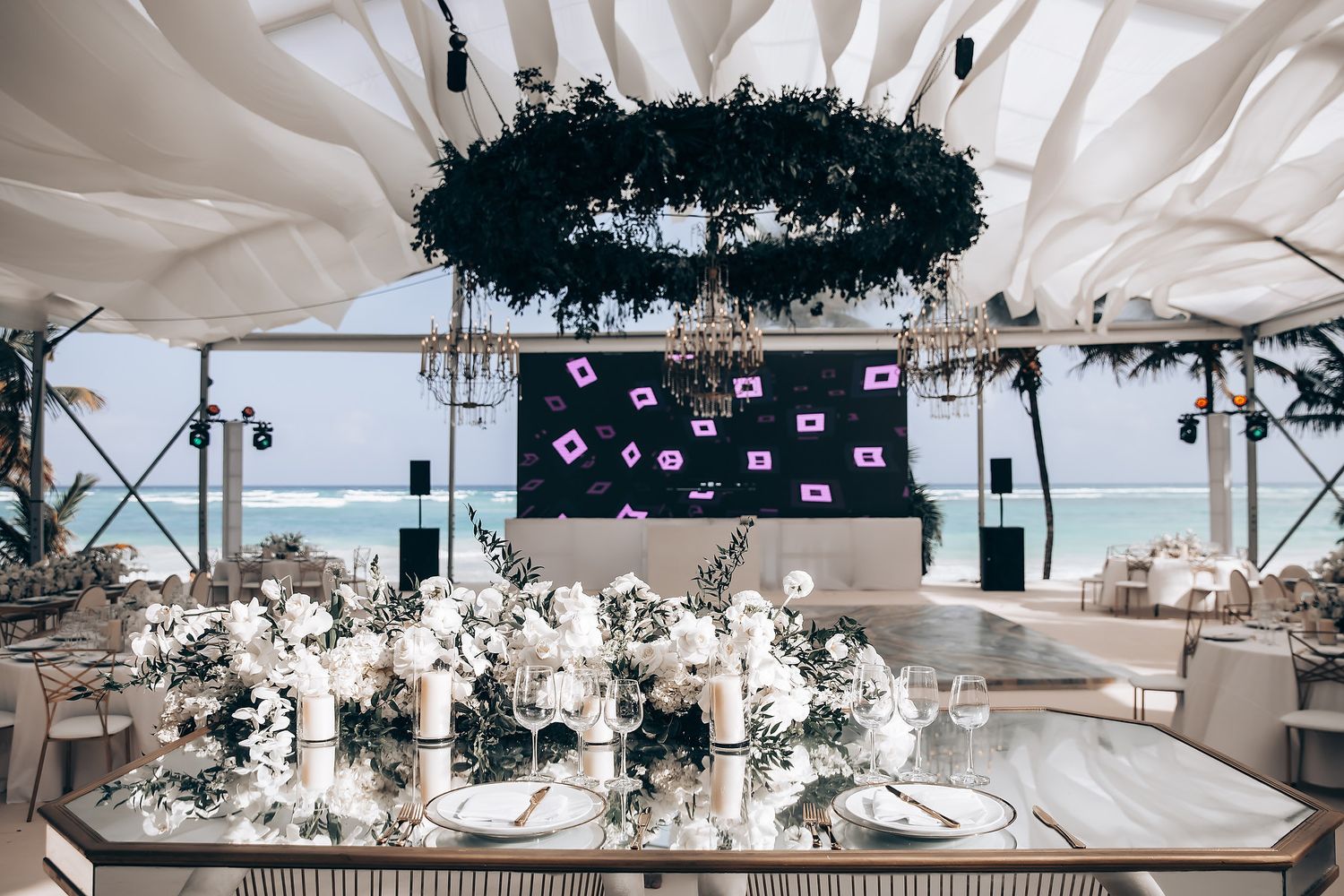 Modern wedding reception with dark floral chandelier, white table settings and a sleek black backdrop.