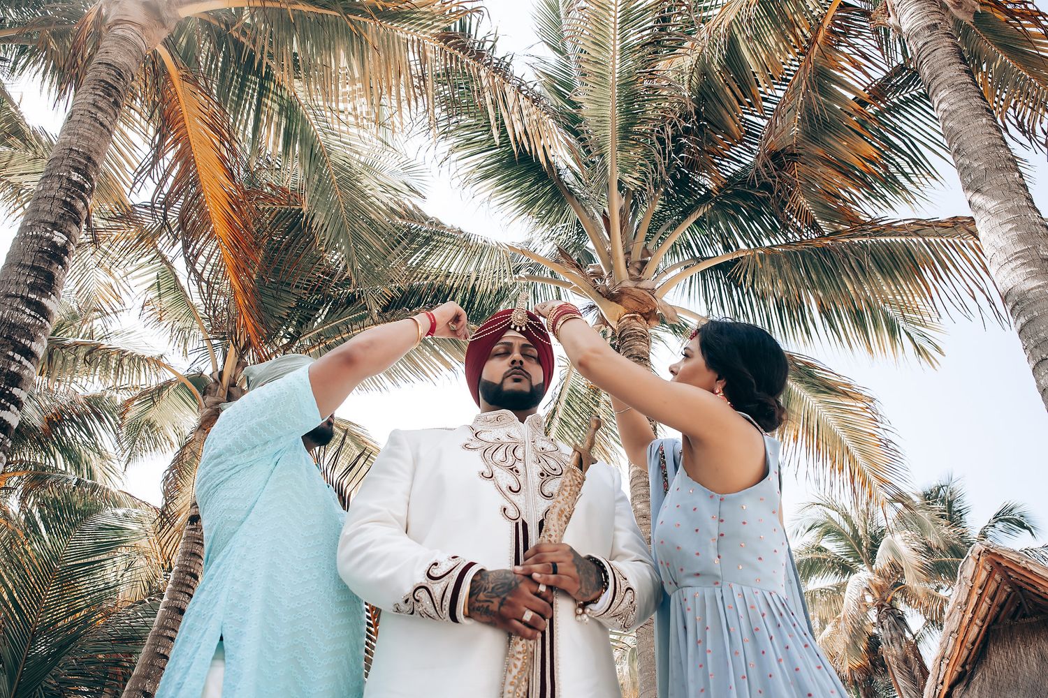 Palm trees frame an outdoor wedding ceremony setting with guests in traditional attire.