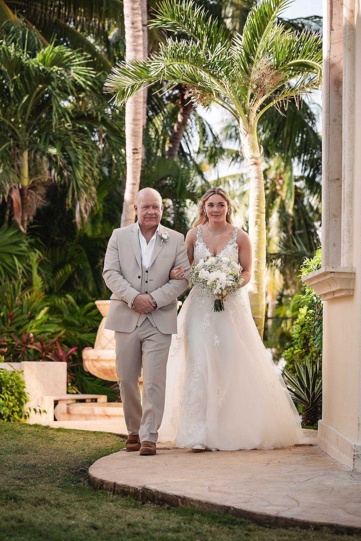 Wedding procession walking under palm-lined colonnade at tropical resort.
