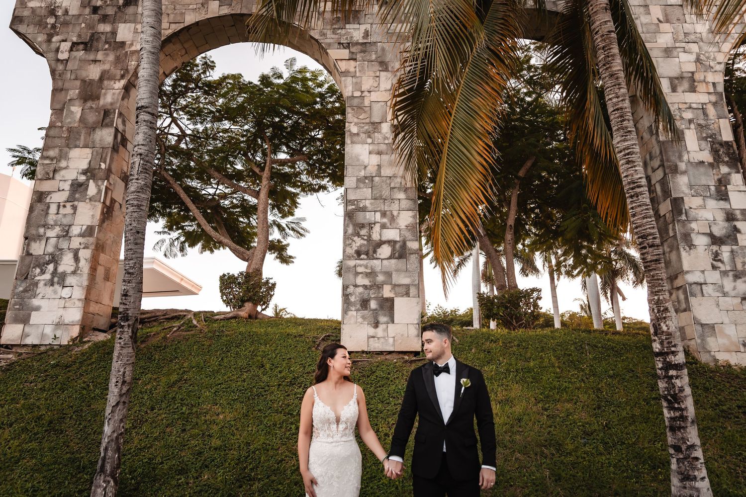 Elegant wedding couple poses beneath historic stone arches with palm trees.