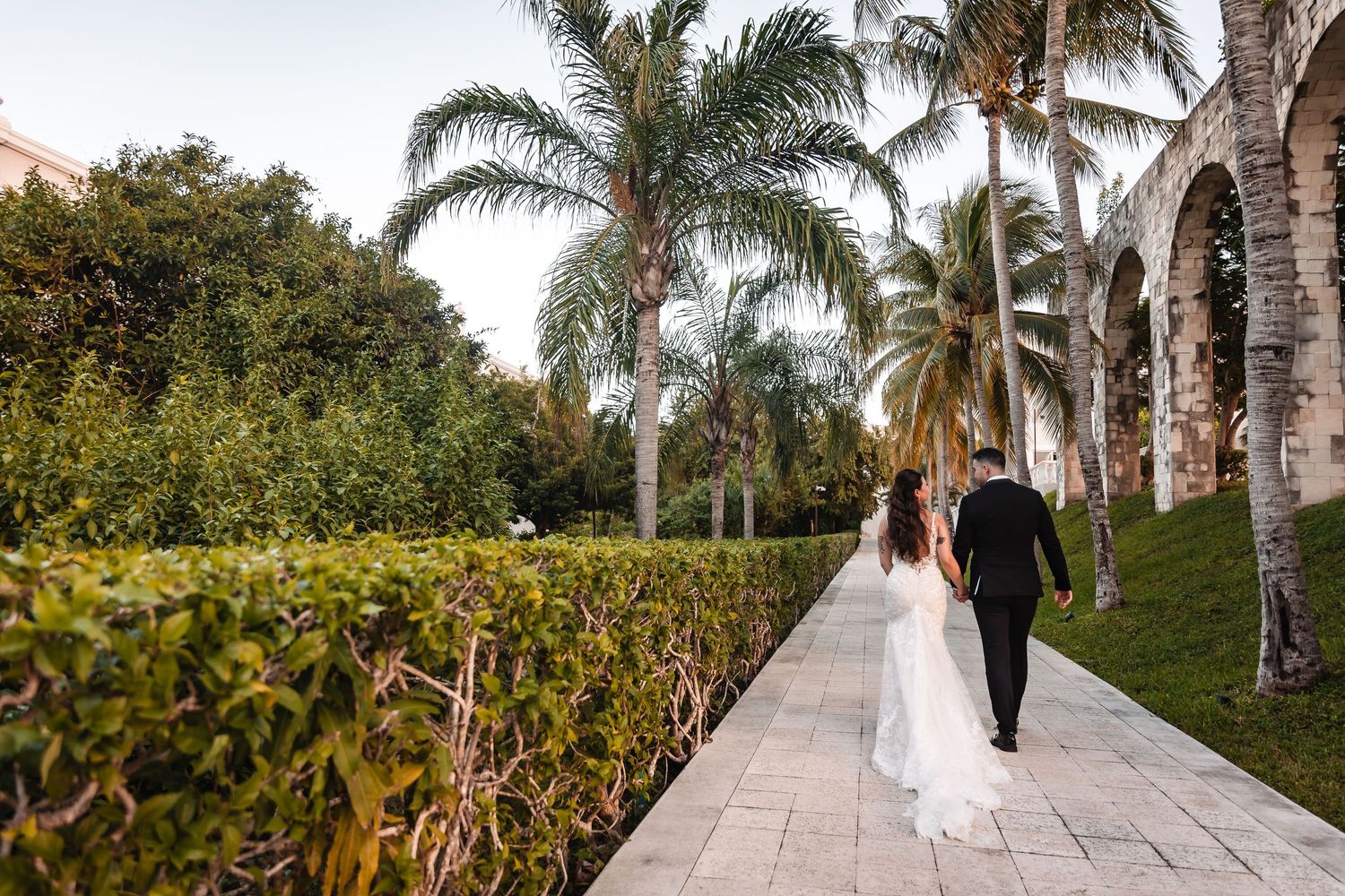 Wedding couple strolls along a scenic palm-lined walkway during golden hour.
