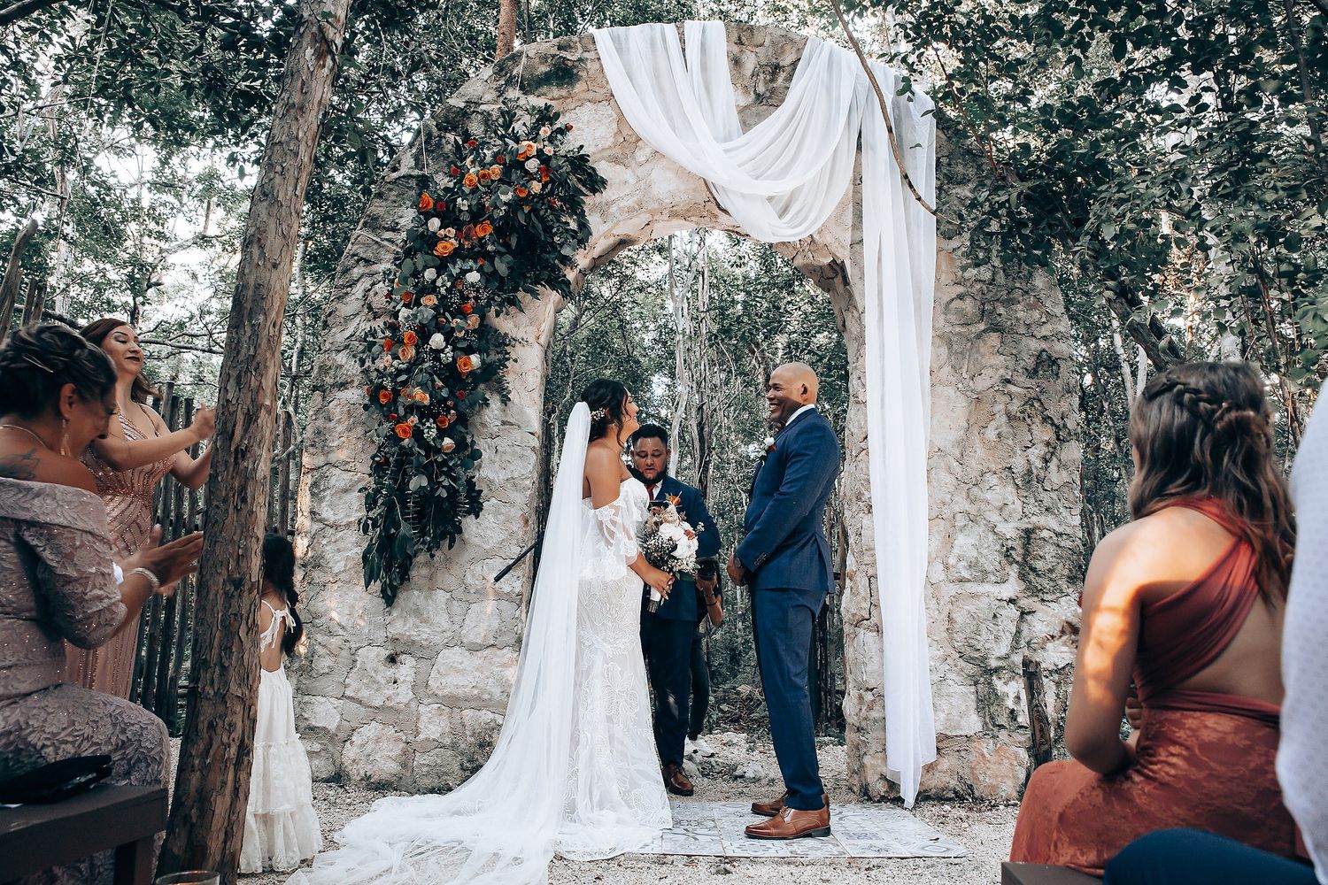 Outdoor wedding ceremony setup in a wooded area with white fabric draped between trees.