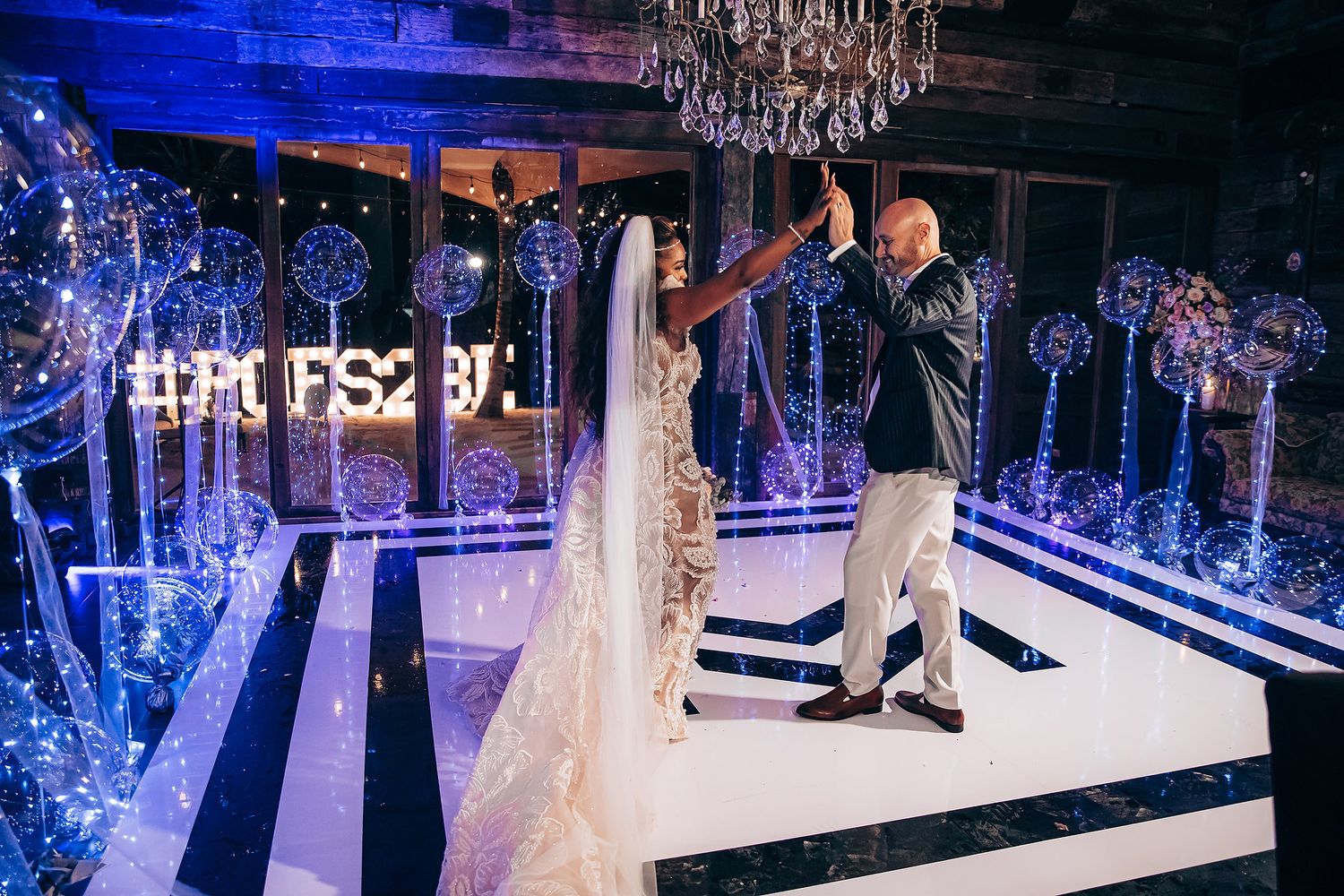 A couple shares their first dance on a black and white striped dance floor illuminated by blue lighting and crystal chandeliers.