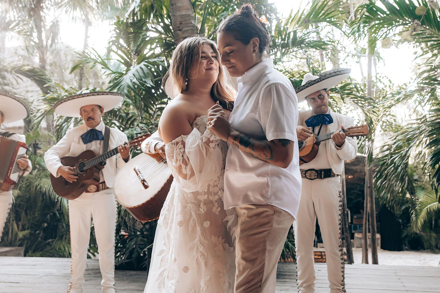 A couple in elegant white attire dances gracefully surrounded by palm trees and tropical foliage at an outdoor venue.