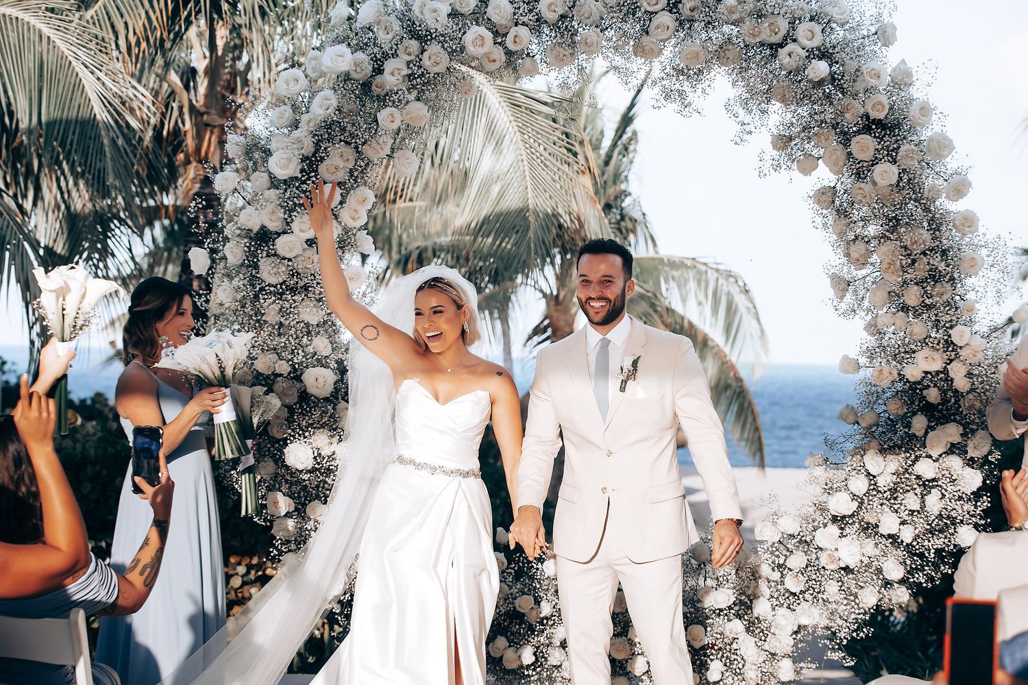 Wedding couple and guests celebrate joyfully under an elegant floral arch by the ocean during sunset ceremony.