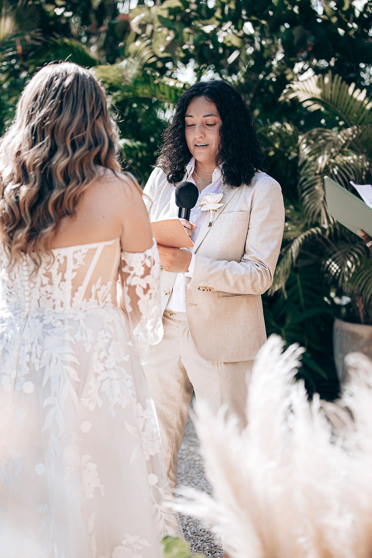 Two women in elegant white outfits share joyful laughter during an outdoor photoshoot with tropical greenery in background.