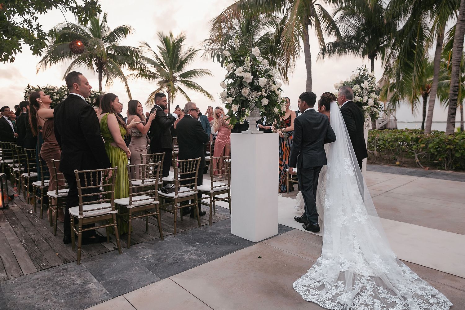 Wedding ceremony guests seated in rows at an outdoor tropical venue with palm trees and ocean views at sunset.