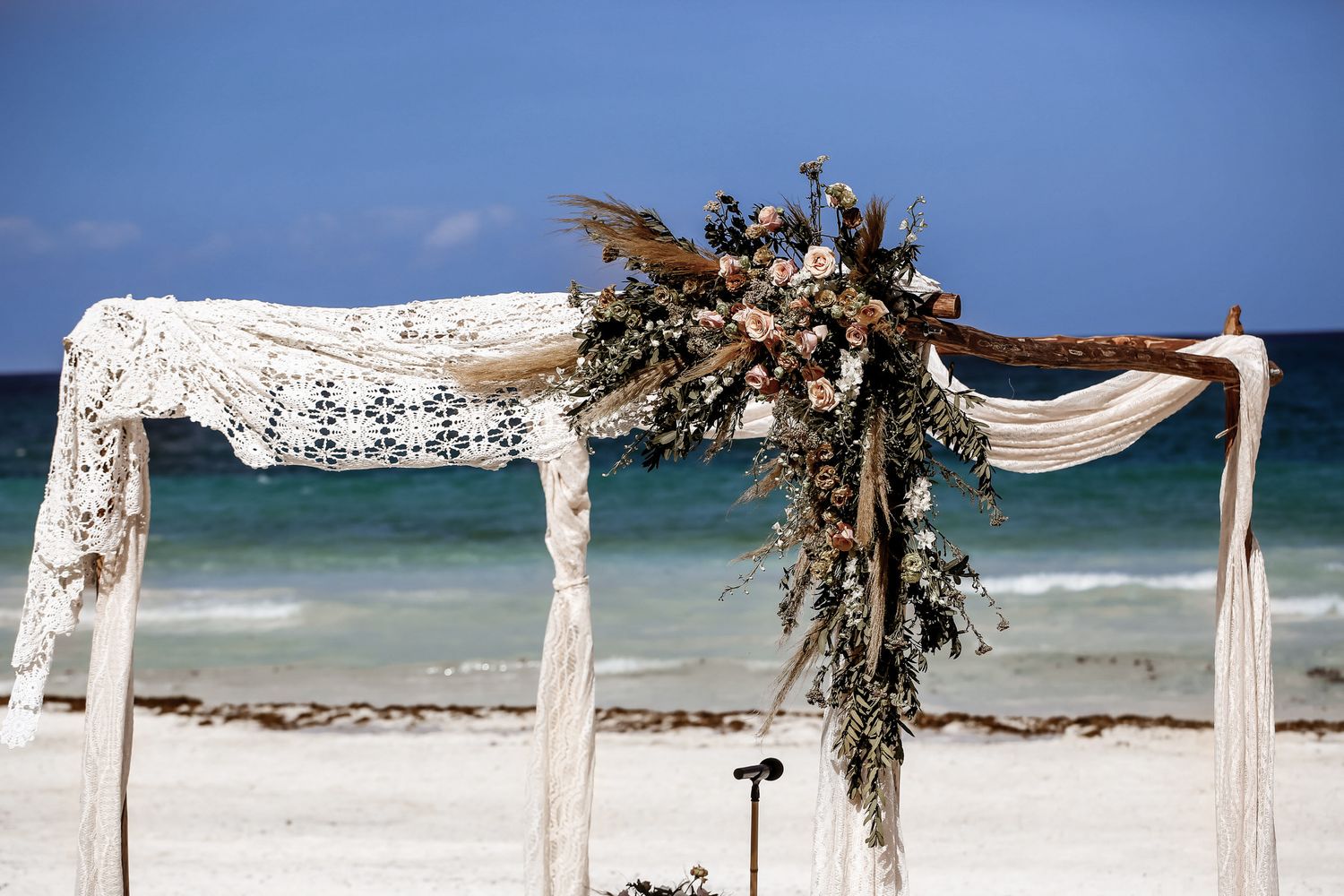 Beach wedding ceremony setup with white fabric draped archway and floral decorations along the shoreline.