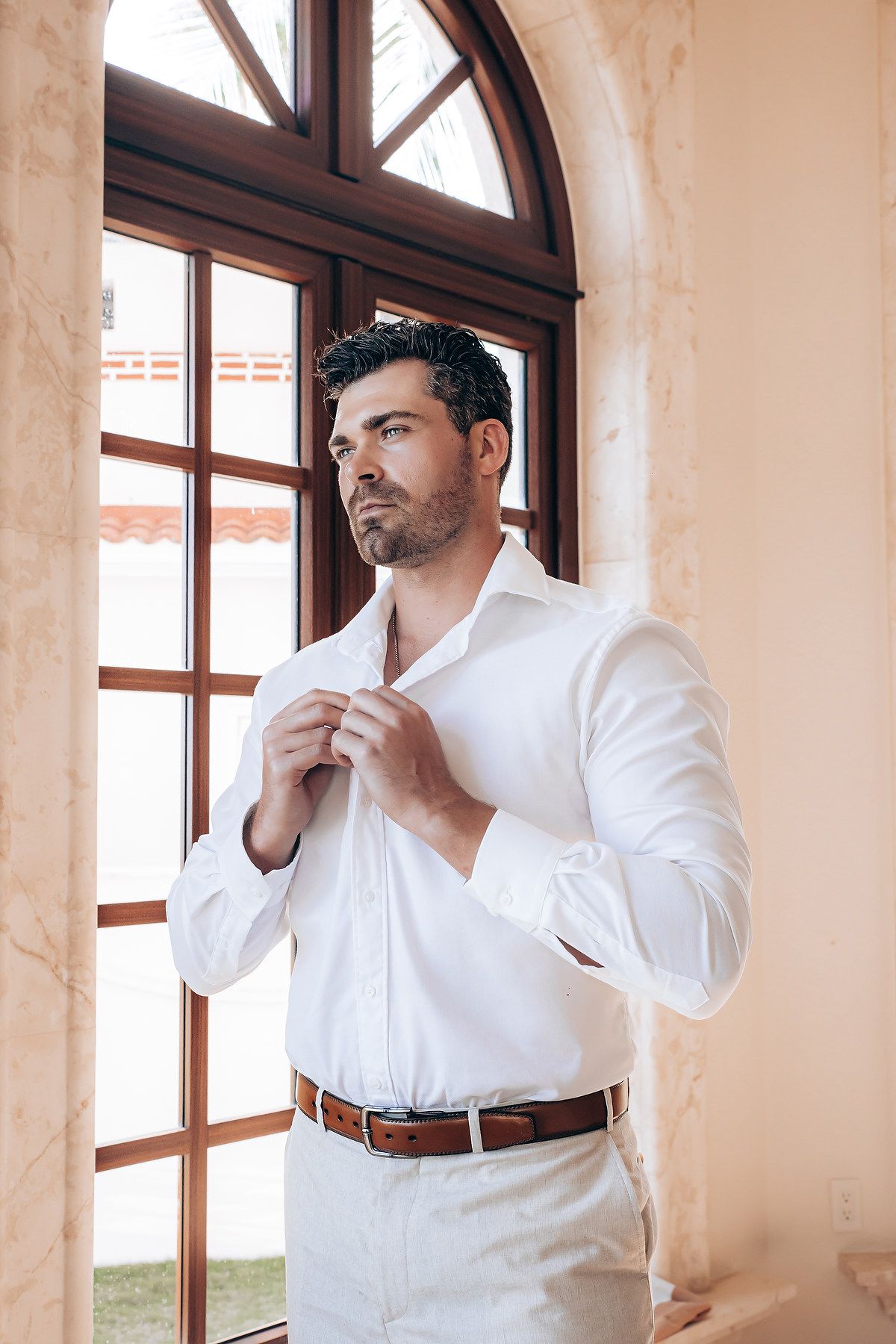Person in white dress shirt adjusting collar while getting ready in a room with arched windows.
