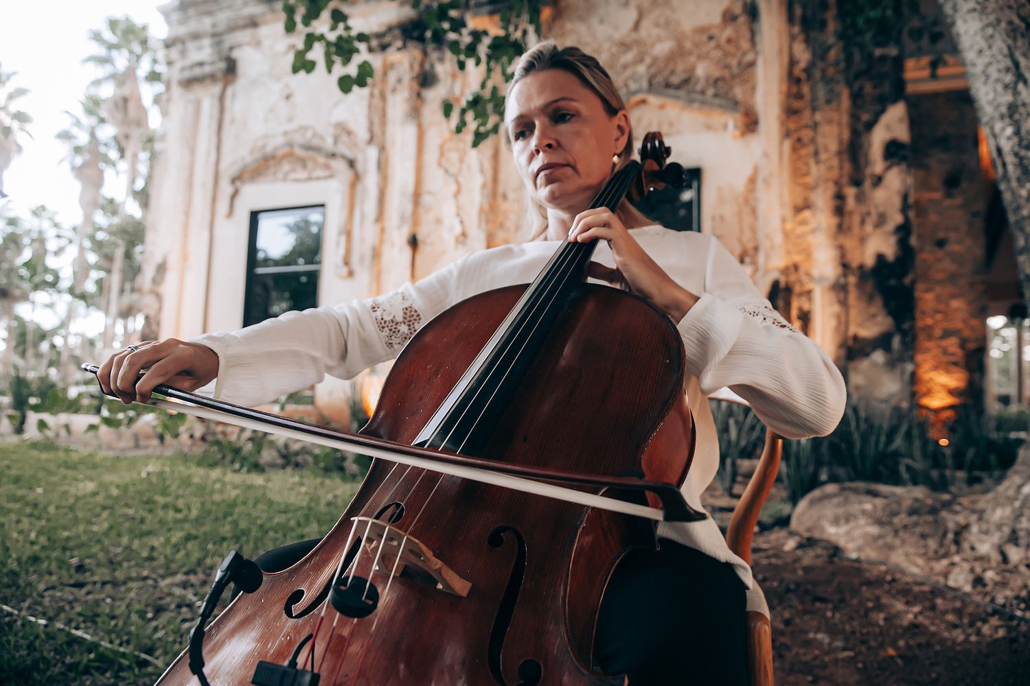 A cellist performs outdoors against a weathered stone wall backdrop with natural lighting and foliage.