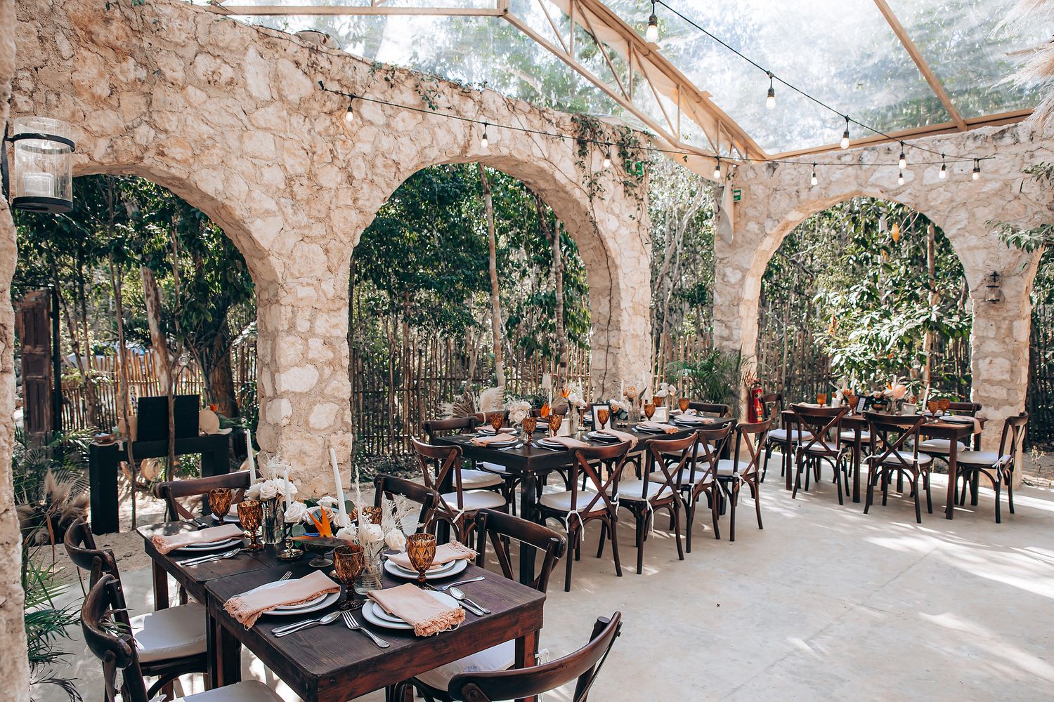 Rustic stone archways and wooden beams create an elegant covered outdoor dining area with tables and chairs.