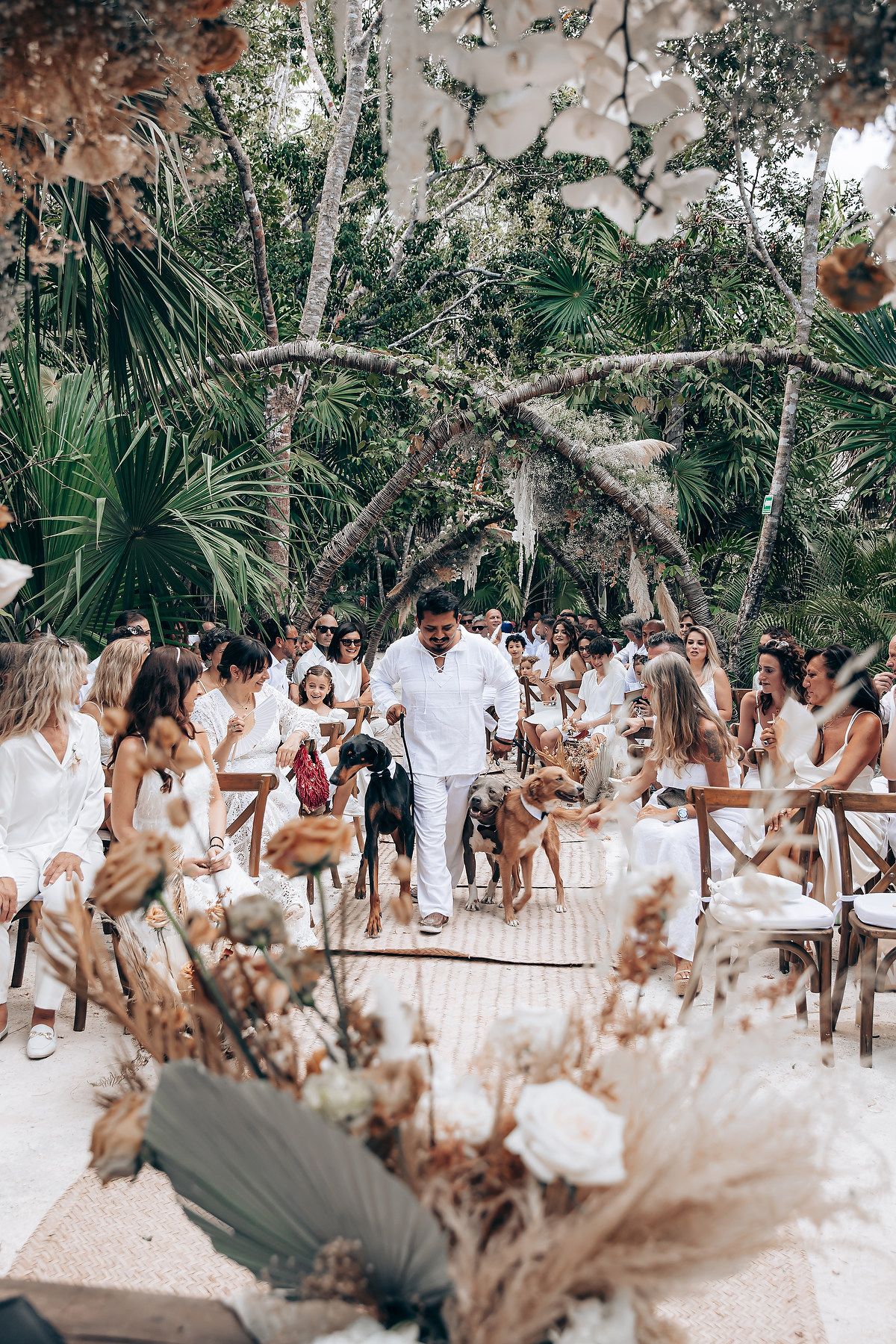 A joyful wedding celebration with guests in white attire dancing together in a tropical outdoor setting with palm trees.