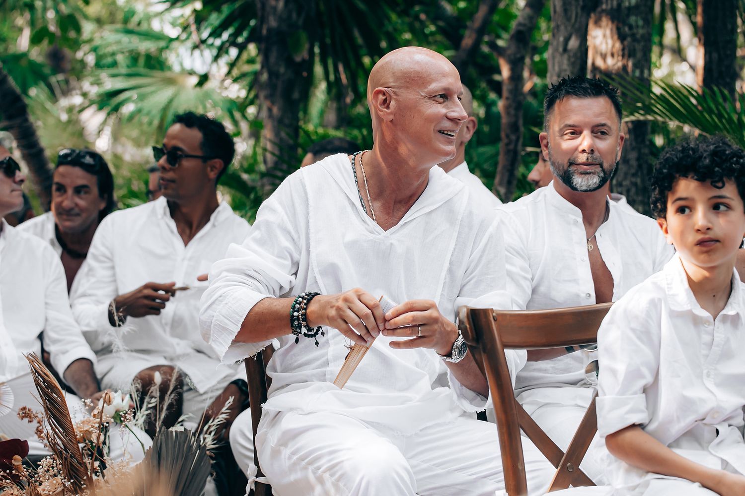 A group of people dressed in white clothing sit together outdoors in a tropical setting.