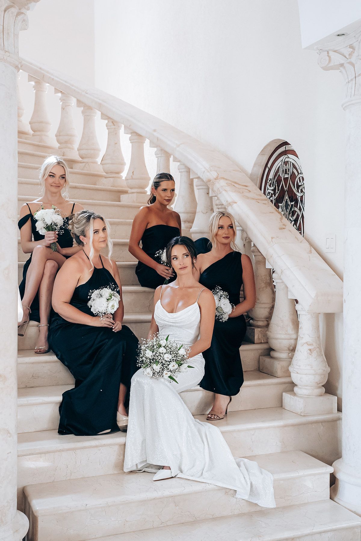 Bridal party in black dresses and white gown arranged on elegant curved marble staircase for formal portrait.