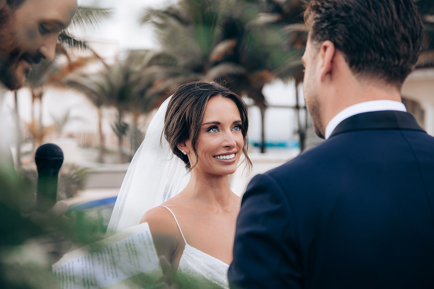 A bride in a white wedding dress smiles joyfully during an outdoor ceremony with palm trees in the background.
