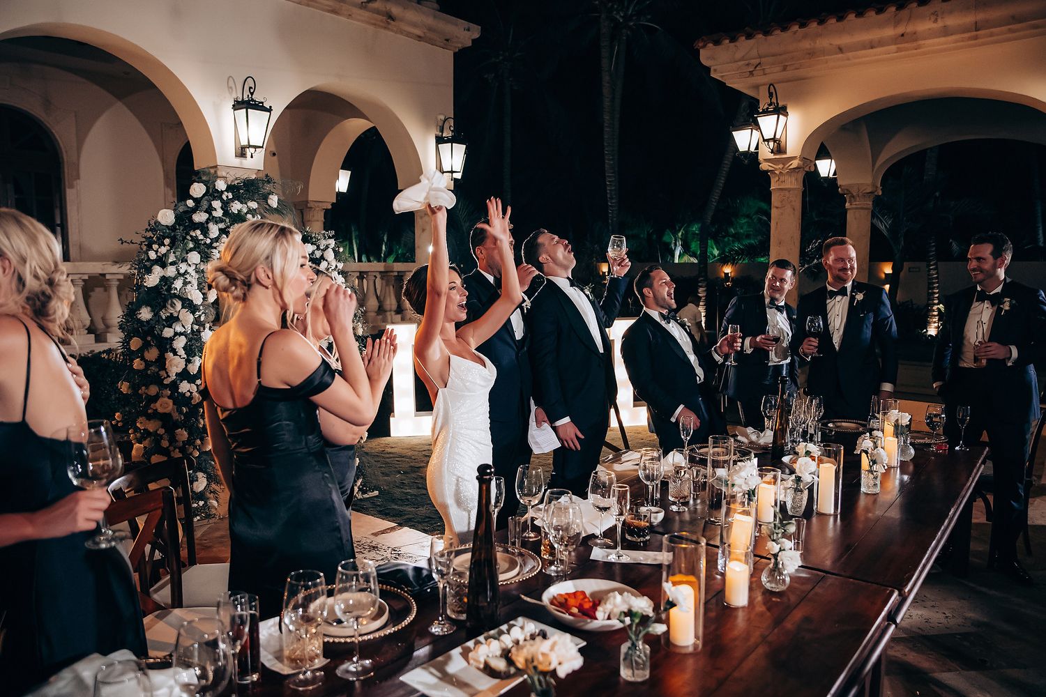 Wedding guests raise their glasses in a toast during an elegant evening reception at a Spanish-style venue.
