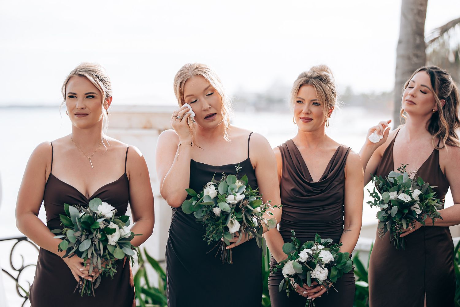 Bridesmaids in elegant black dresses laugh together while holding green and white bouquets on a sunny day.
