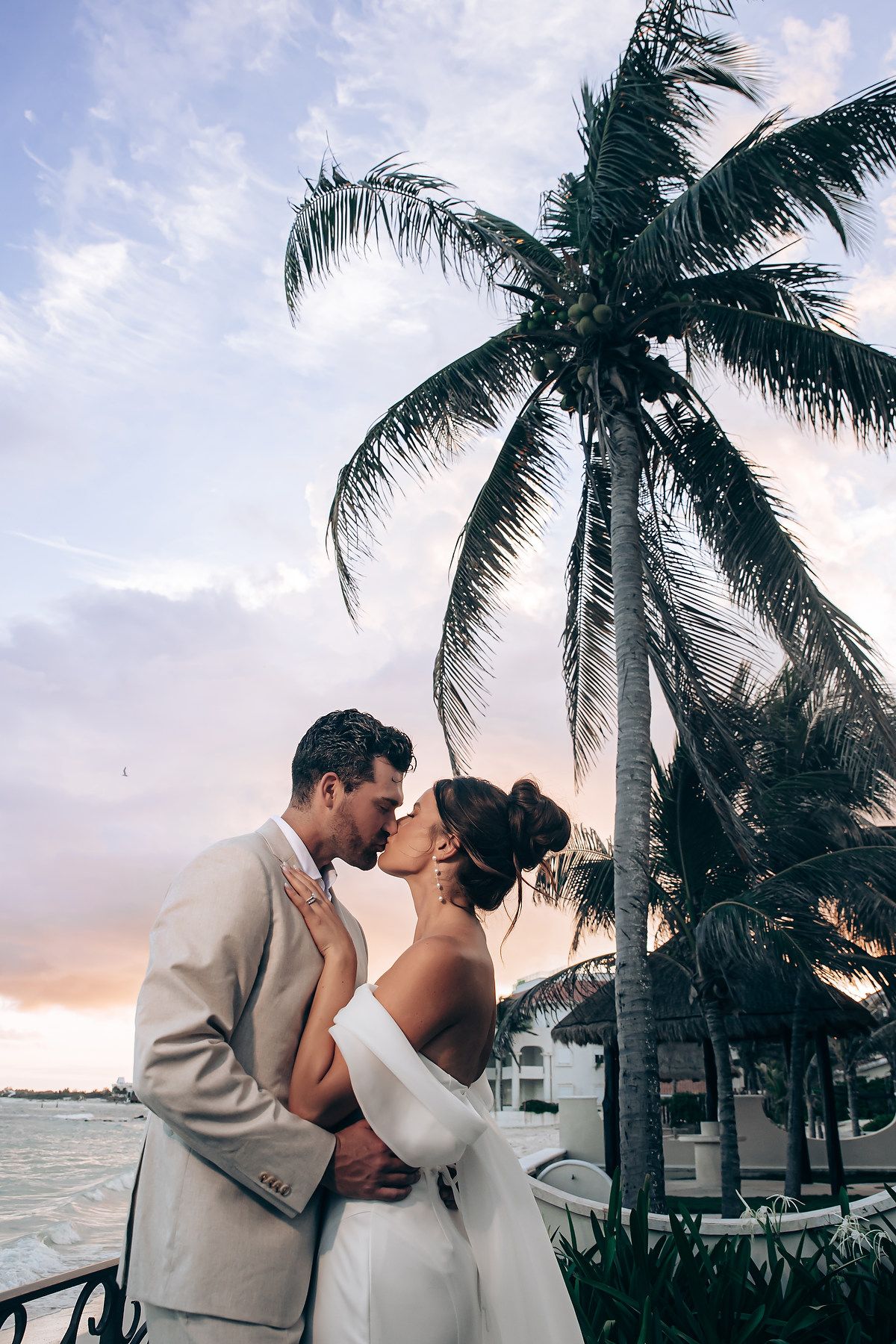 A romantic couple embraces and kisses on a tropical beach at sunset with palm trees swaying in the background.