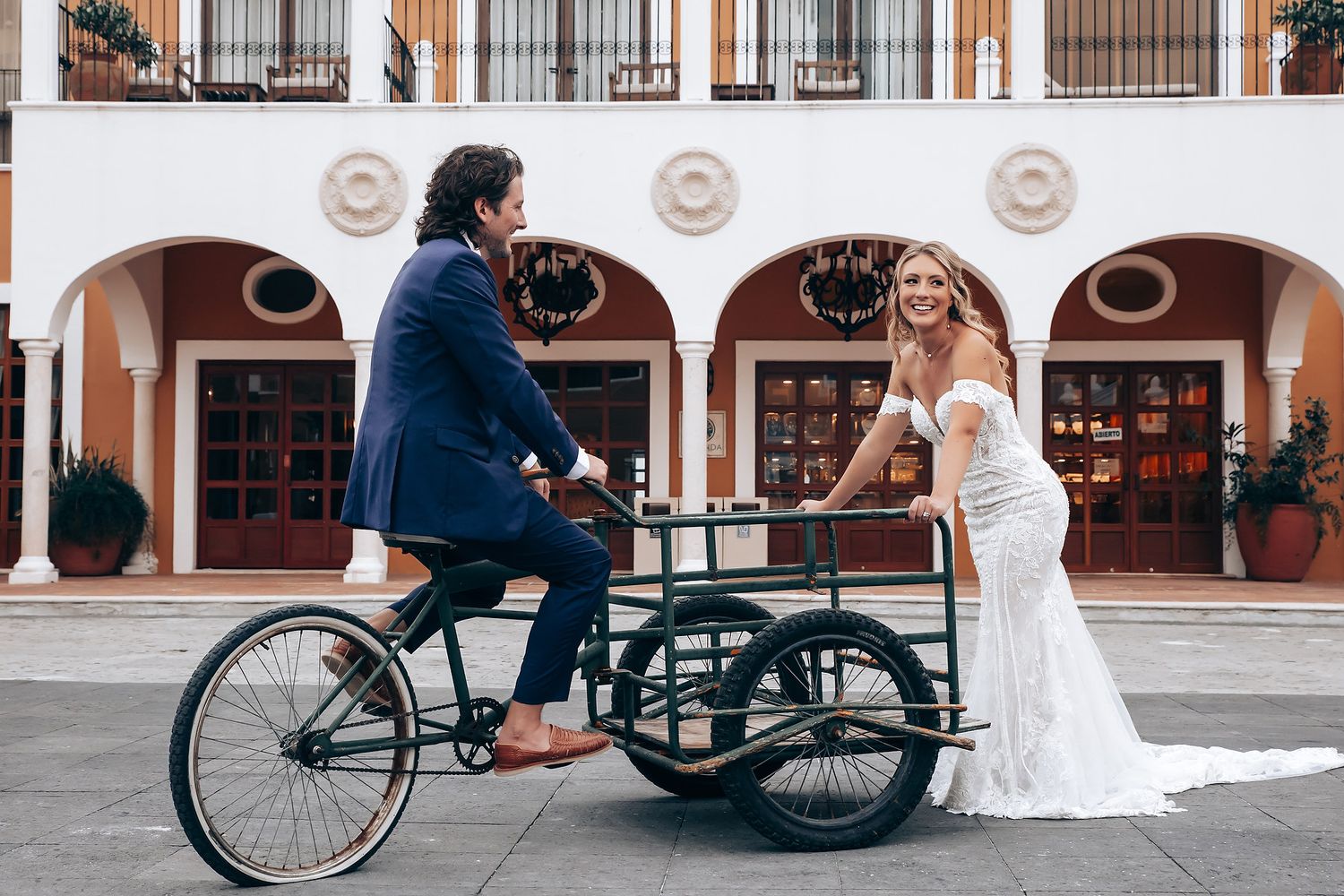A couple poses playfully with a vintage bicycle in front of a white Spanish-style building.
