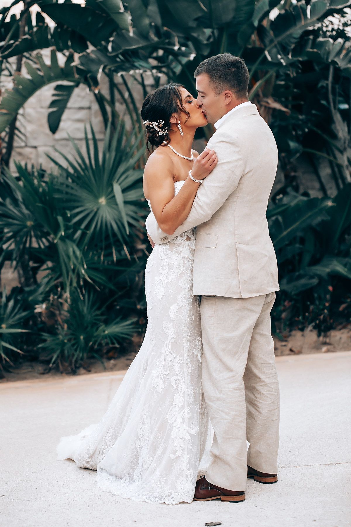 A romantic wedding couple shares an intimate moment on a beach path surrounded by tropical plants.