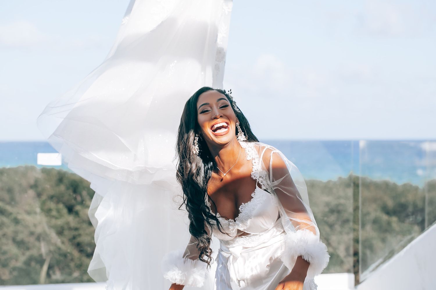 A beaming bride in a white dress poses joyfully against a bright outdoor backdrop.