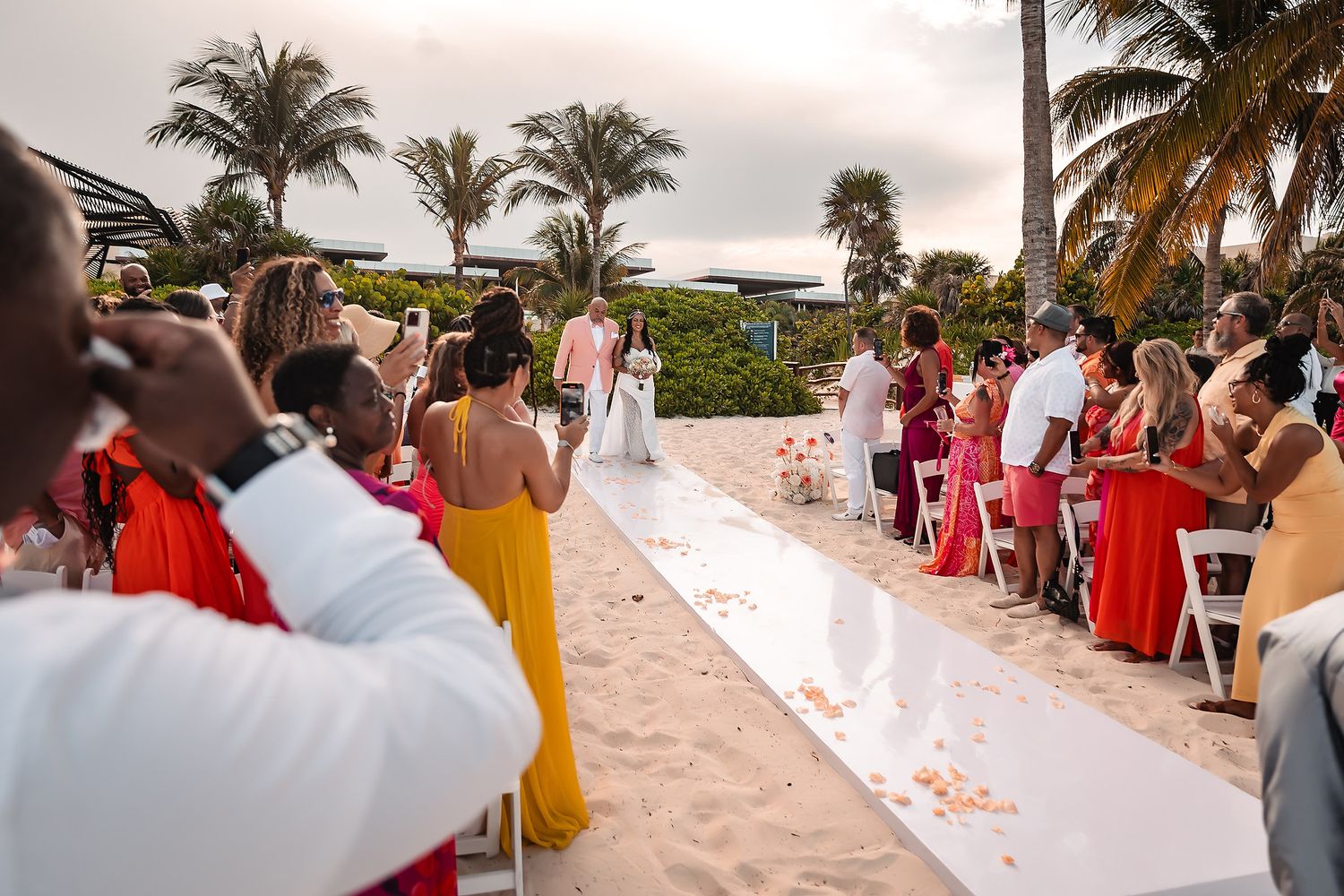 Beach wedding ceremony setup with guests seated in chairs on sand near palm trees.