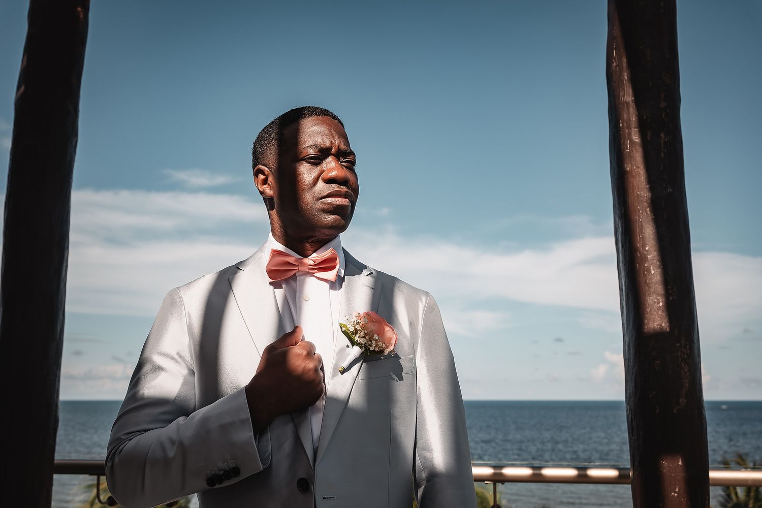 Groomsmen in formal attire stand against oceanfront backdrop on sunny day.