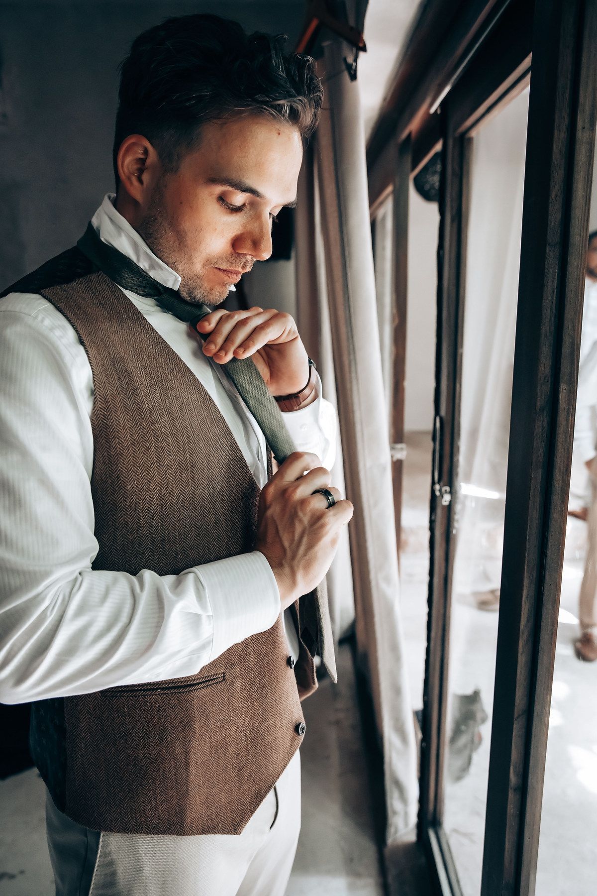 Person in brown vest and white shirt standing thoughtfully by window.