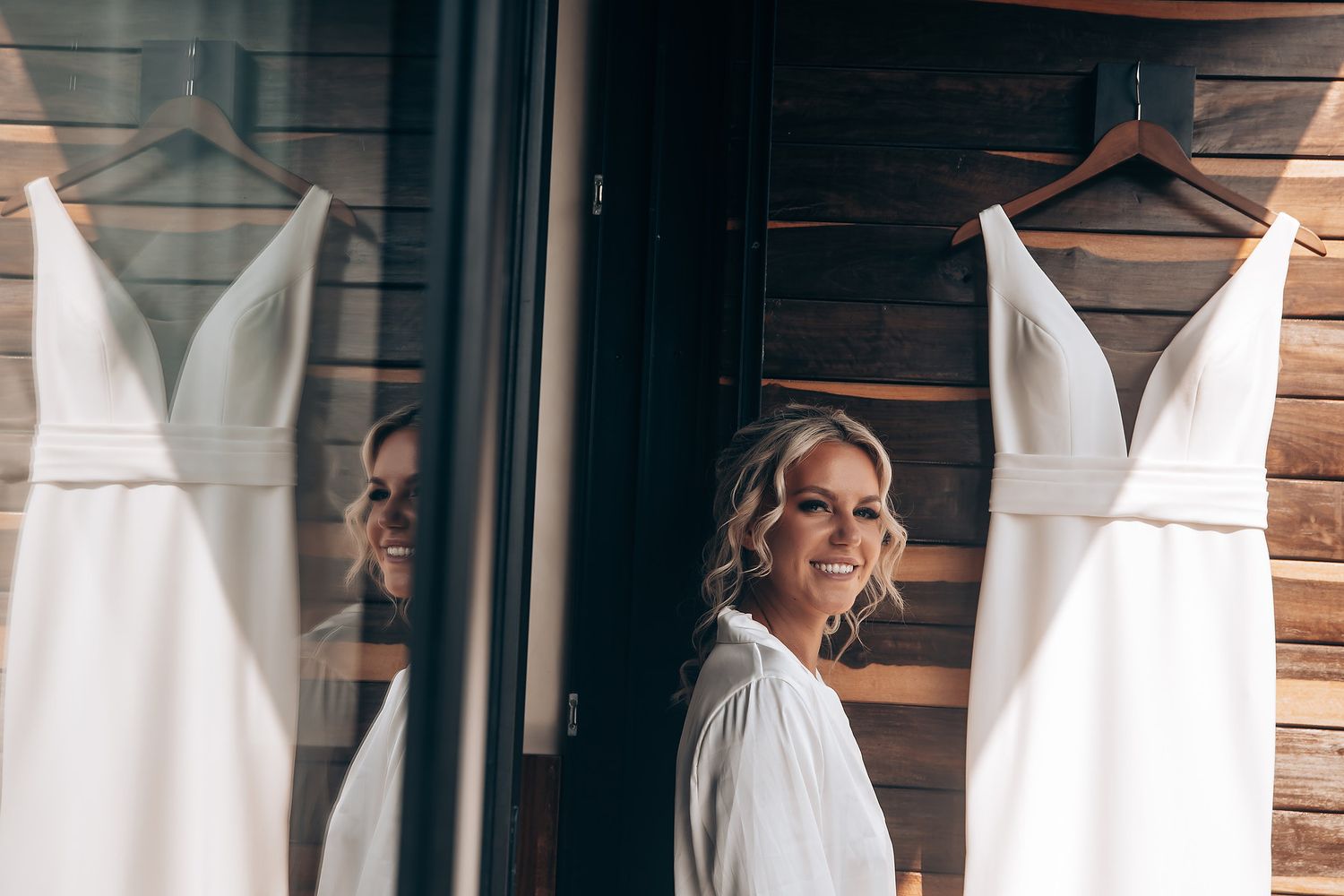 Wedding dress hanging on rustic wooden wall with mirror reflection.
