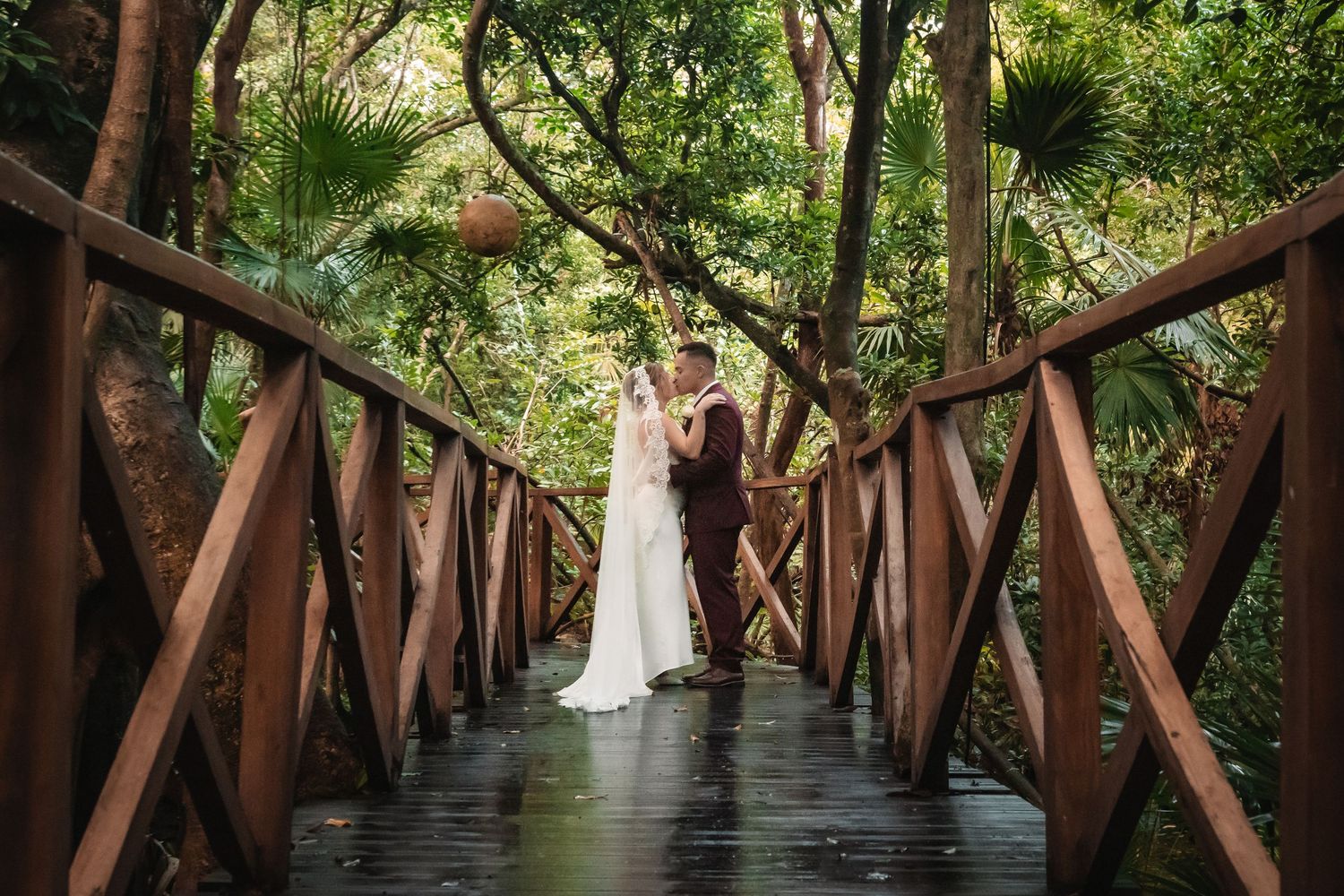 Wedding photo on wooden bridge walkway surrounded by lush greenery.
