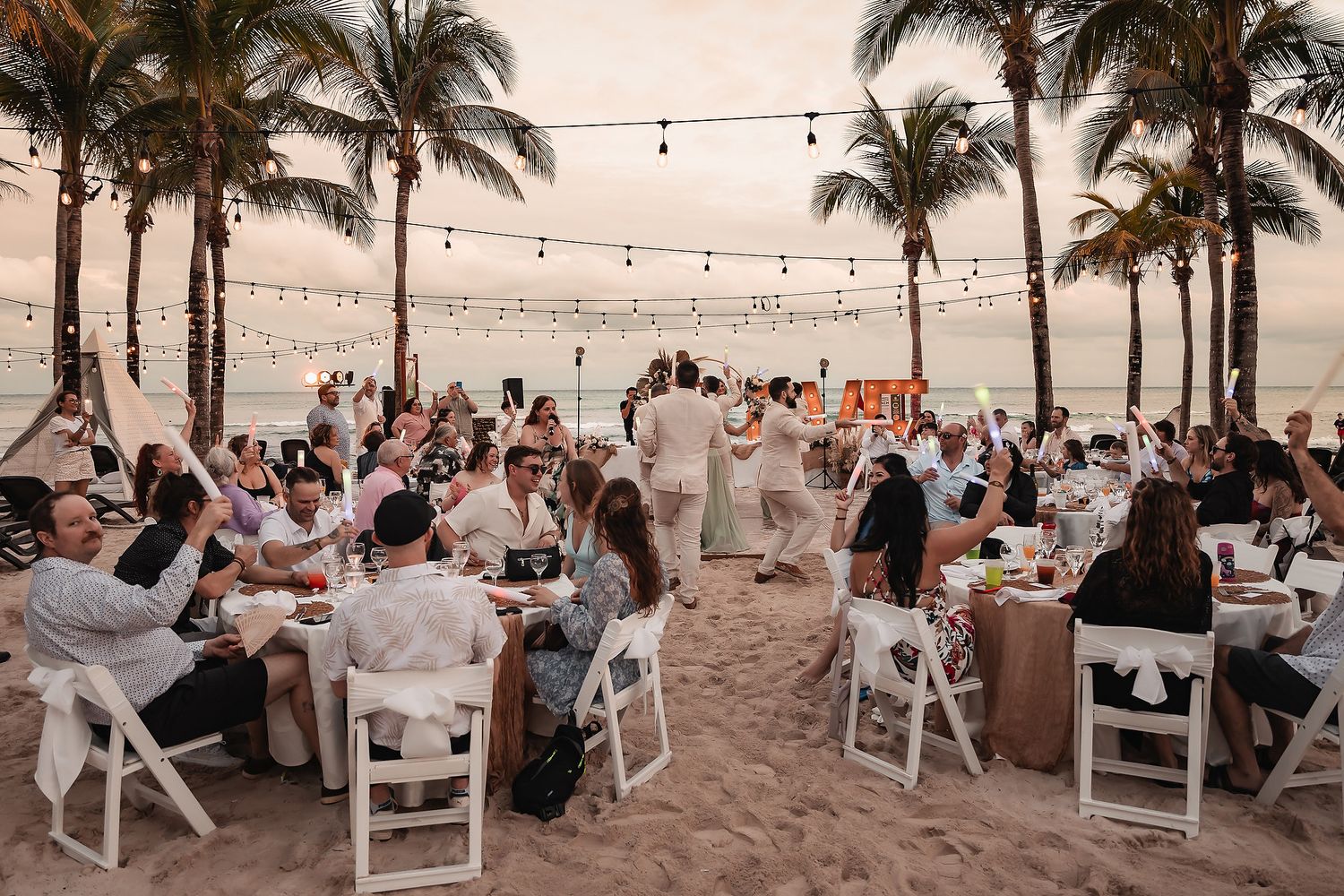 Outdoor beach wedding reception with string lights and guests seated at white tables.
