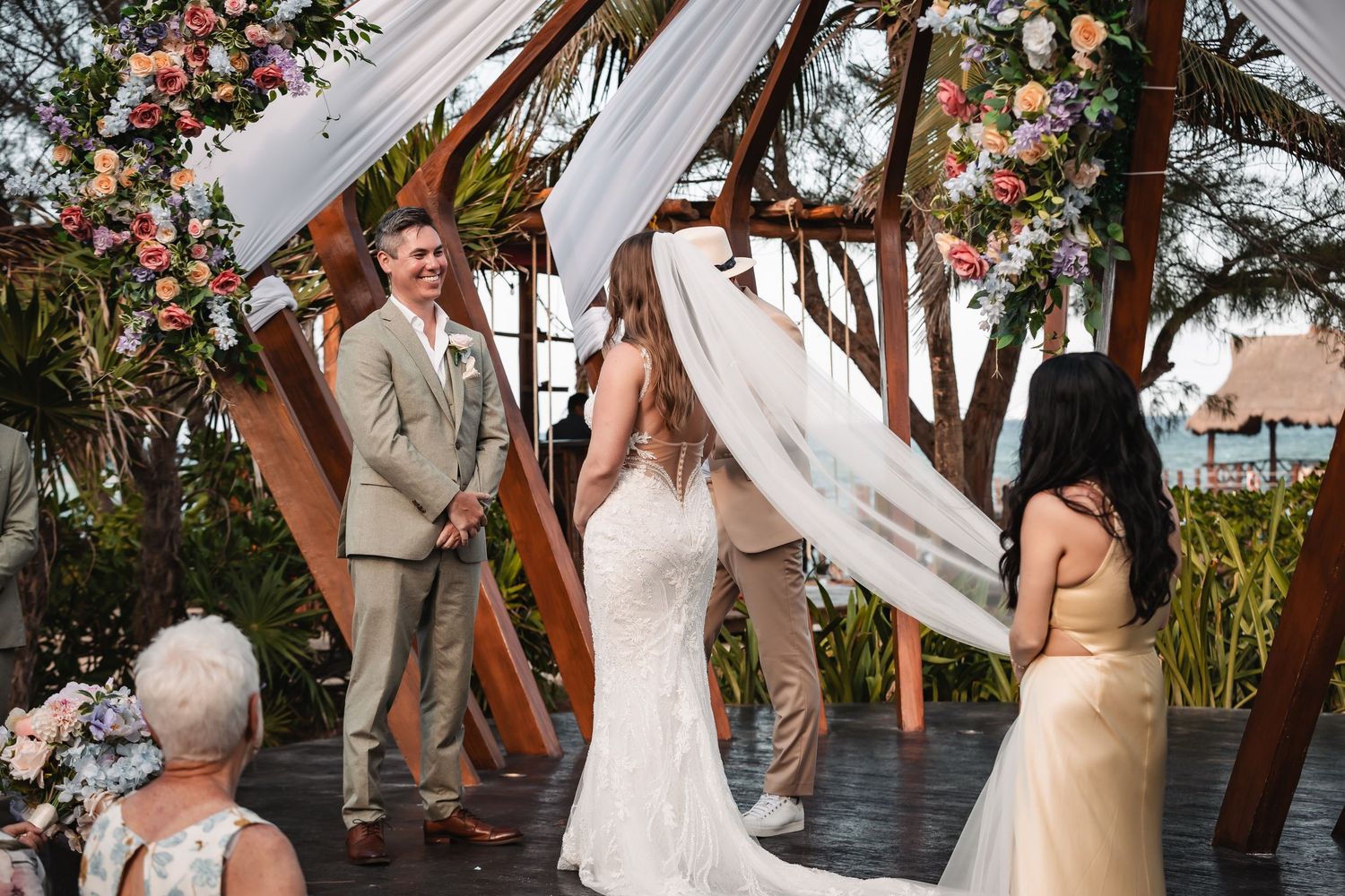 Intimate beach wedding celebration under wooden arch decorated with white fabric and floral arrangements.