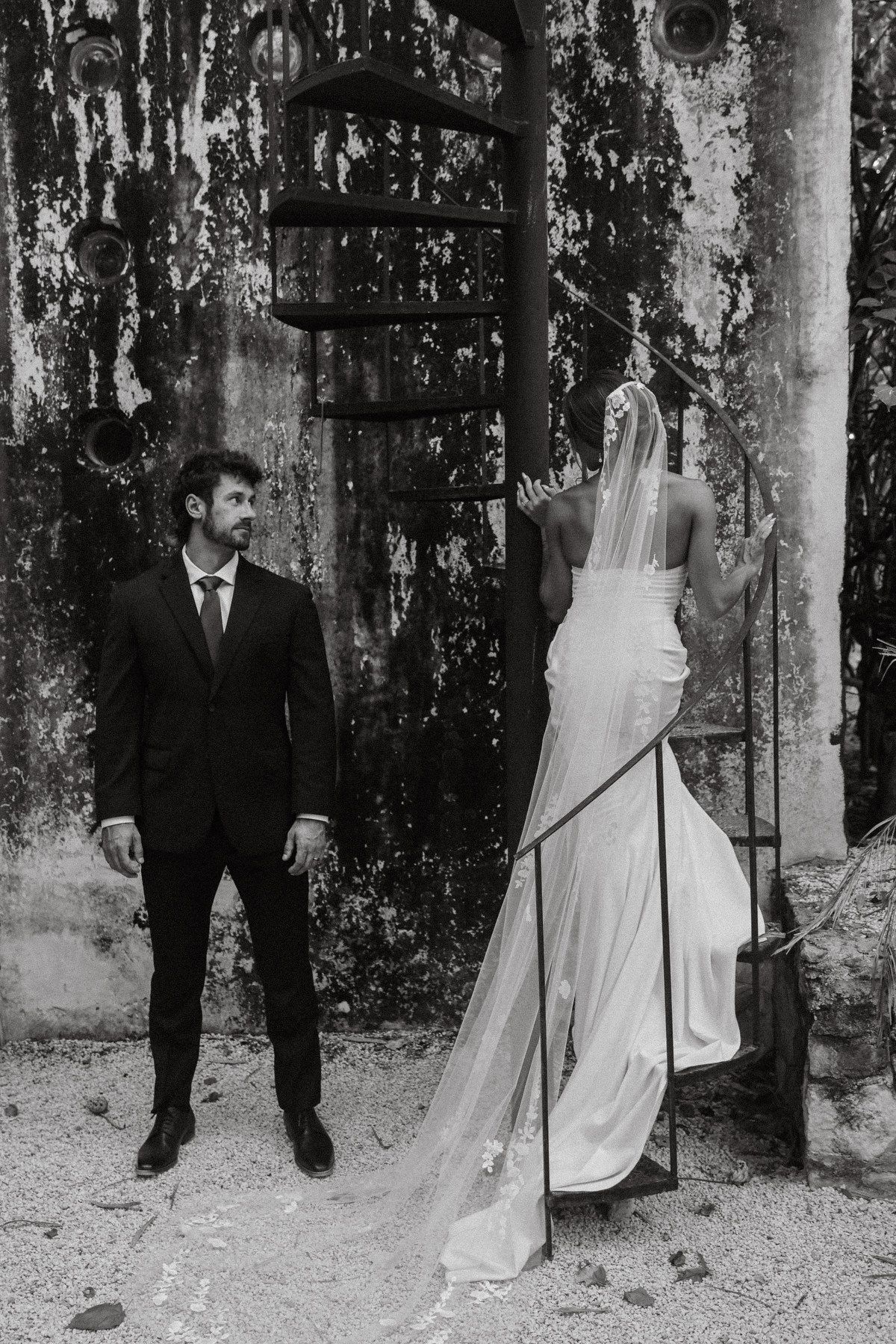 Black and white wedding photo of groom in suit watching bride in backless gown climb ornate iron staircase.