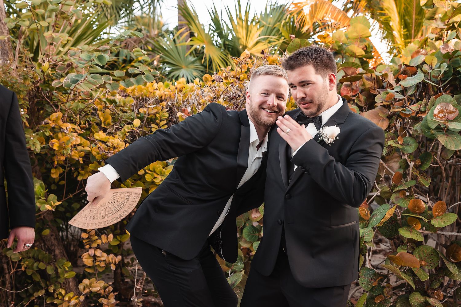 Two groomsmen in black tuxedos laugh and pose playfully outdoors among lush tropical greenery at a wedding.