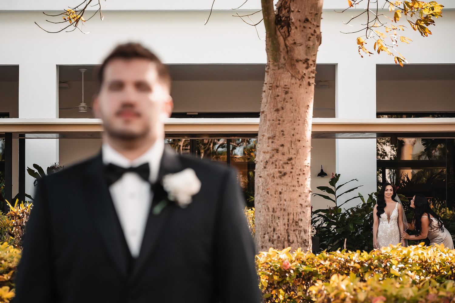 Groom in a black tuxedo stands in the foreground as the bride waits behind a tree in a lush outdoor setting.