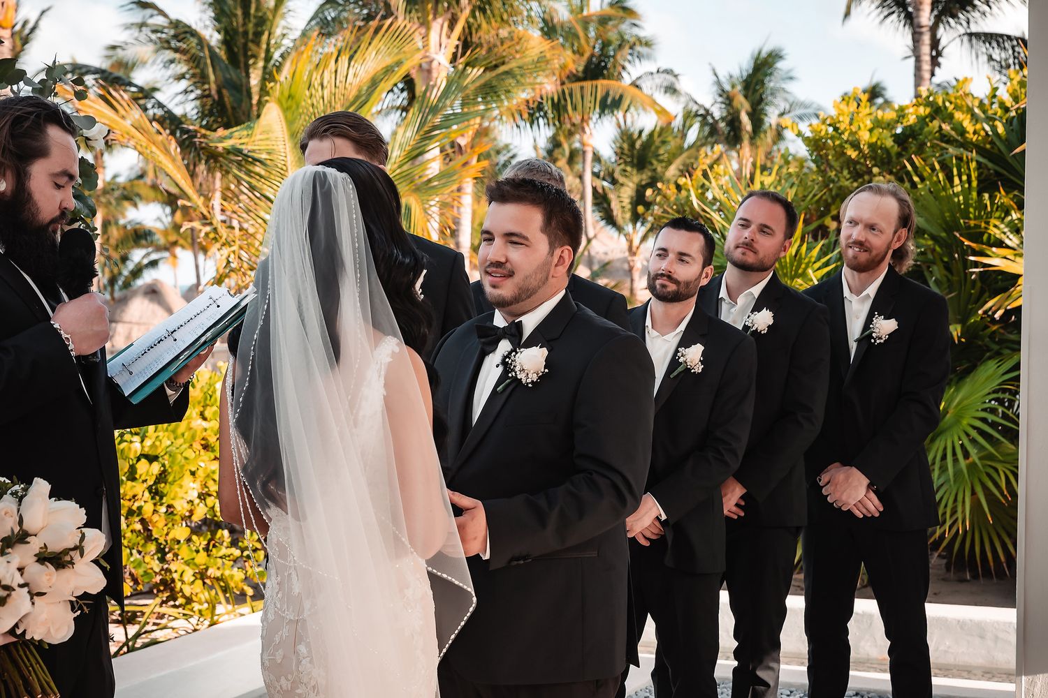 Groom smiling at bride during outdoor wedding ceremony with groomsmen in black suits and tropical palm trees backdrop.