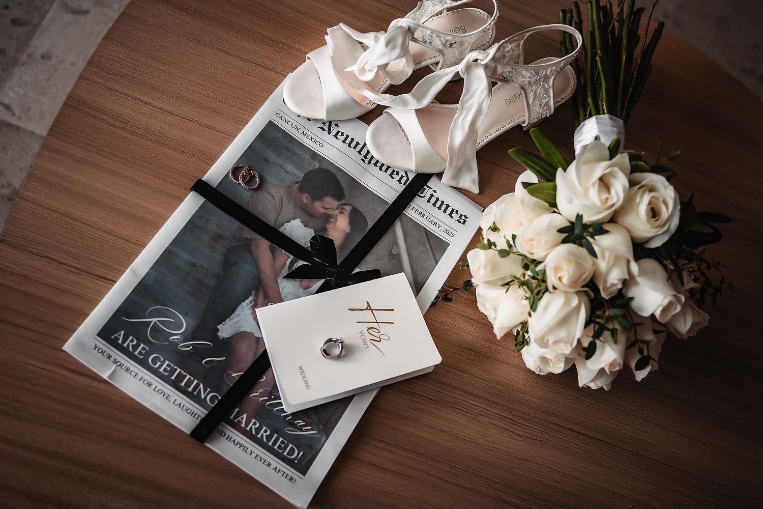 Wedding flat lay with white roses bouquet, bridal heels, engagement ring, and newspaper announcement on wood table.