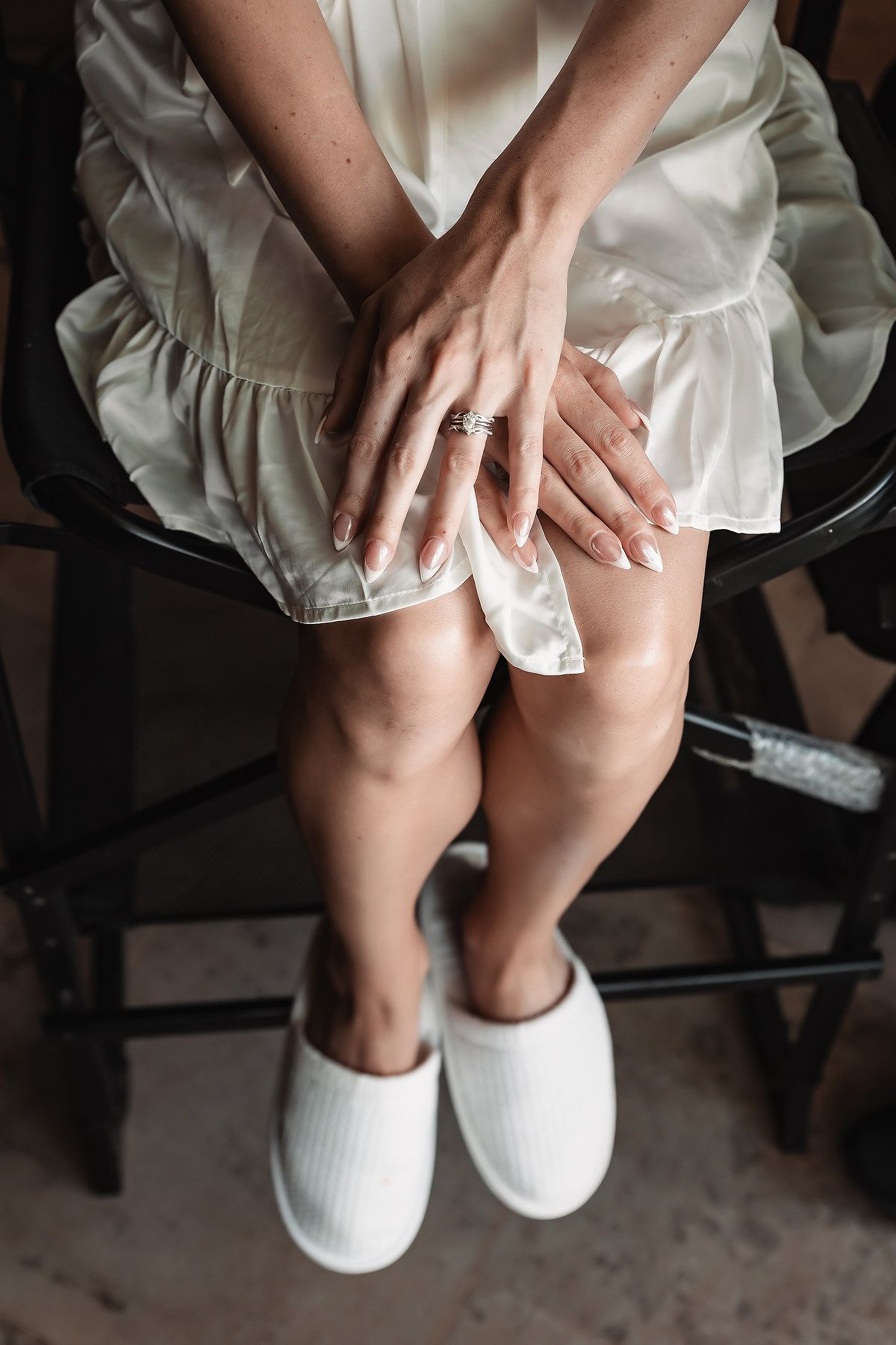 Bride in white silk robe sitting on chair, showing engagement ring and French manicure, wearing white slippers.