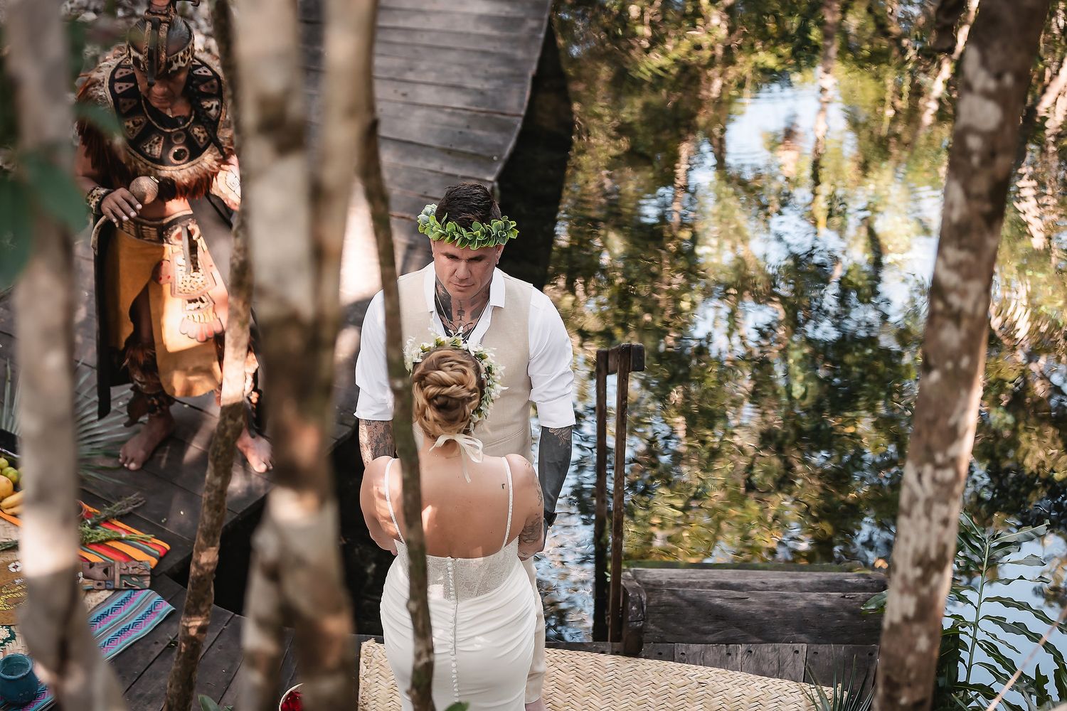 Bride and groom share intimate moment on wooden steps surrounded by trees near tranquil water.