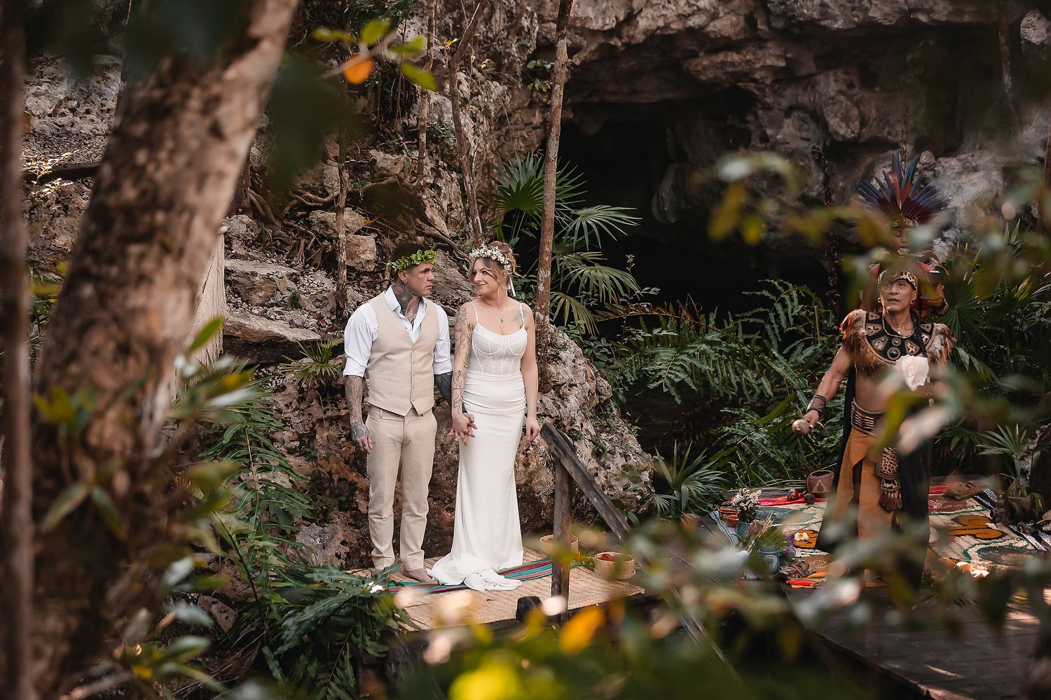 Couple exchanges vows in lush jungle cave ceremony with officiant in traditional attire and colorful floral altar.