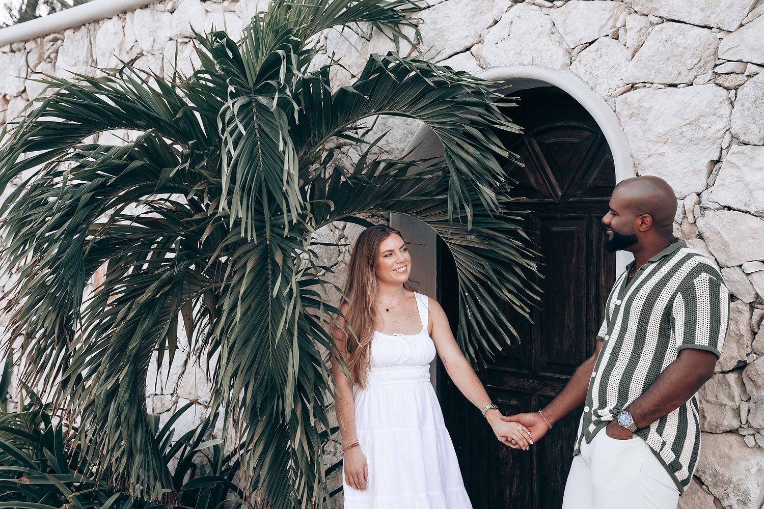 Interracial couple holding hands smiling at each other near a stone arch with tropical palm.