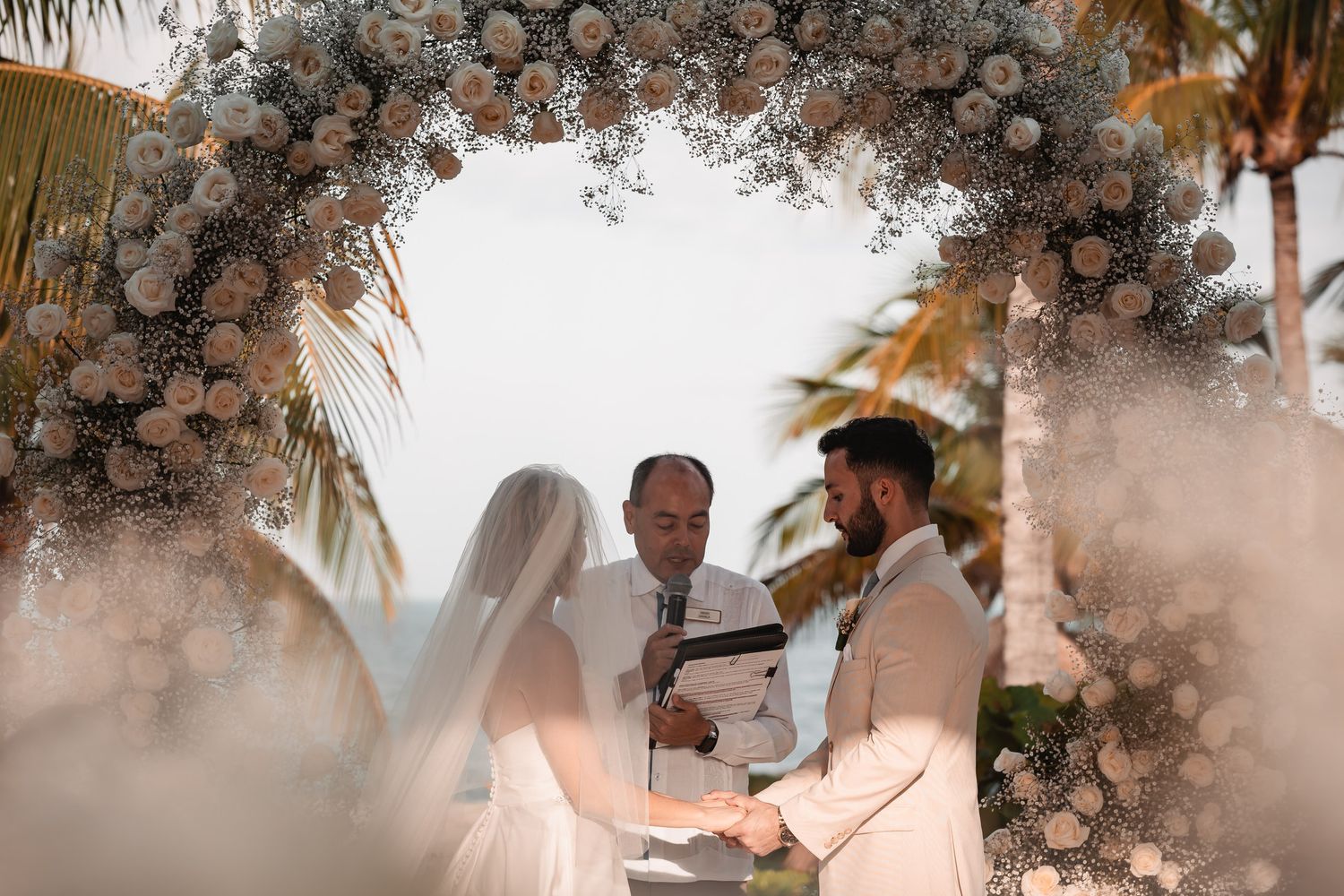 Beach wedding ceremony under floral arch with officiant and couple.