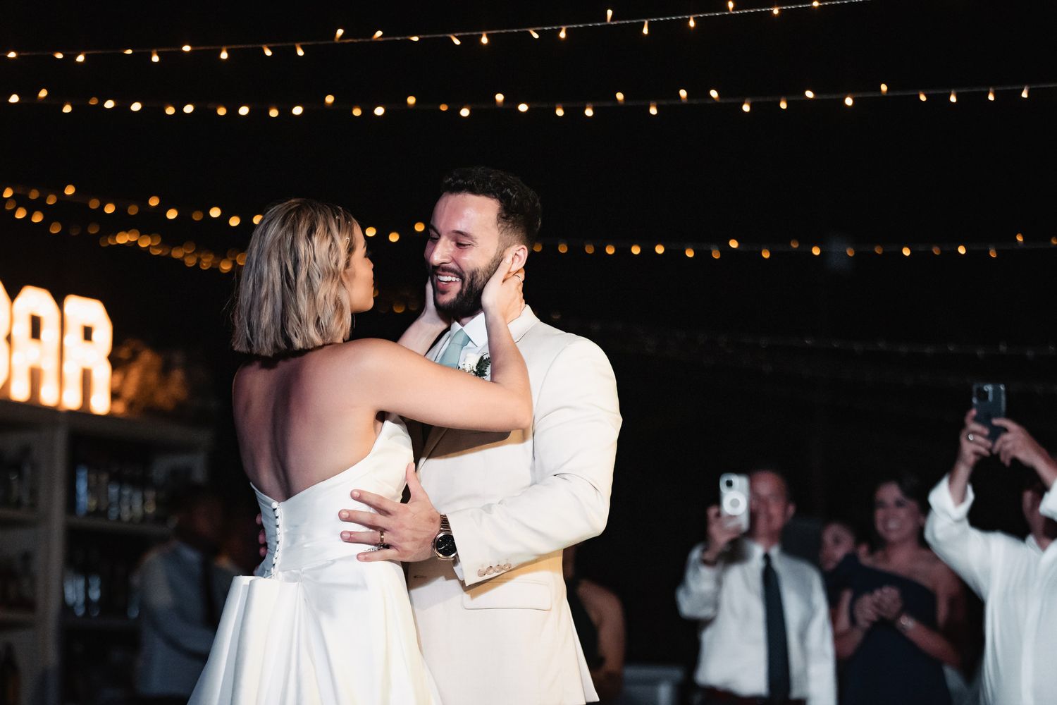 Bride and groom share first dance under warm string lights at wedding reception.